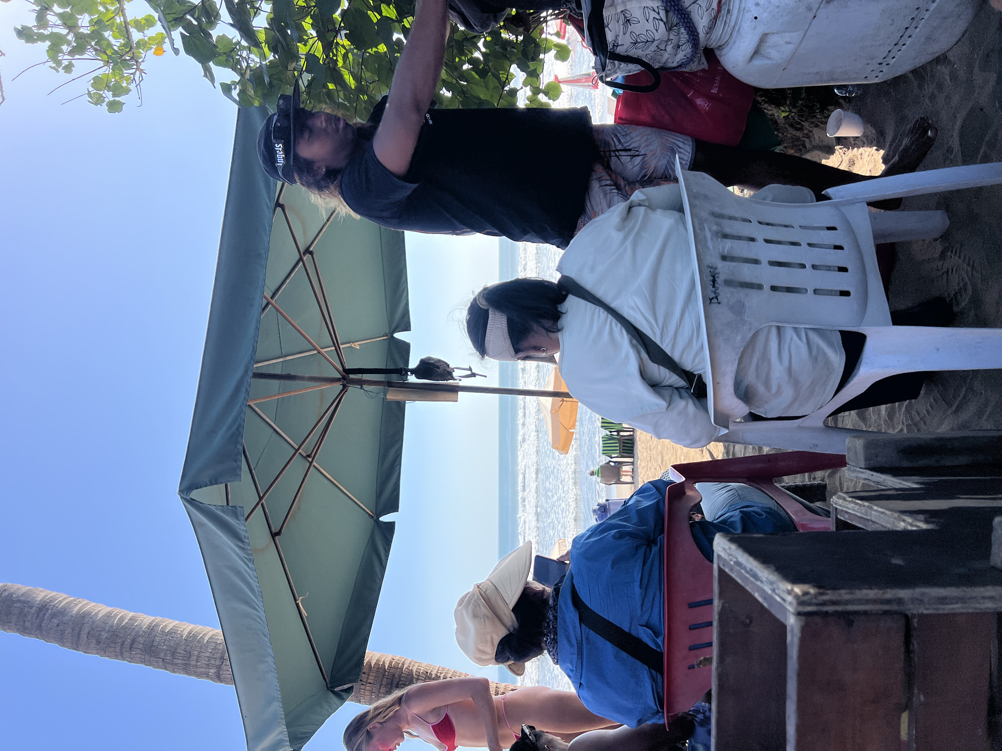 The image depicts a beach scene in Kuta, Indonesia, under a clear blue sky. A large, teal-colored umbrella provides shade for several individuals. In the foreground, a person wearing a blue shirt and a light-colored hat is seated, looking at their phone. To their right, another person in a white jacket is also seated. A man with long hair, wearing a black t-shirt and patterned shorts, stands near the umbrella, appearing to be interacting with someone or something out of frame. To the left, a woman in a pink bikini stands near a palm tree. The background shows the ocean with gentle waves and a few beach chairs. The ground is covered in sand.