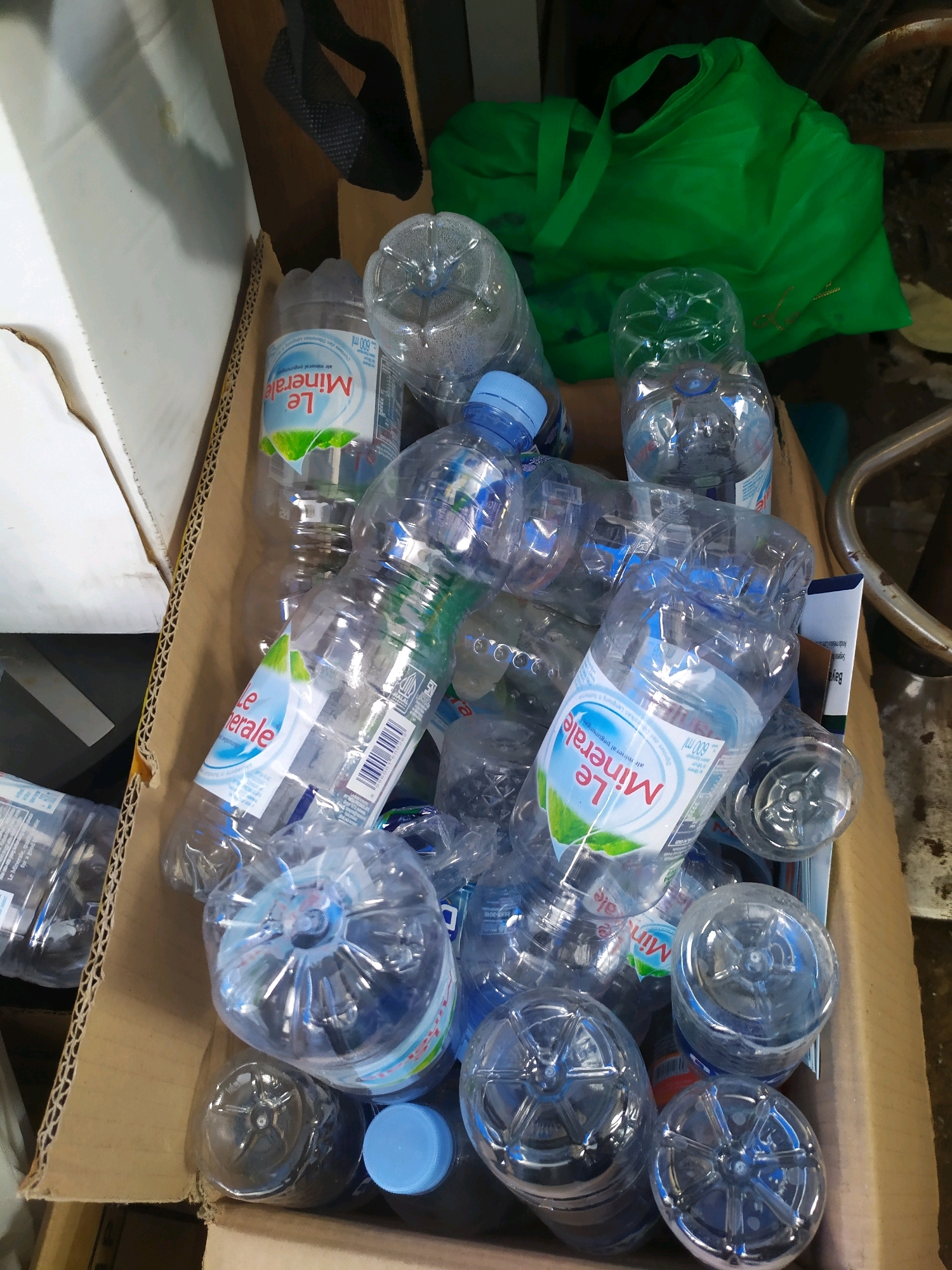 A close-up, overhead shot shows a cardboard box filled with an assortment of empty plastic water bottles. The bottles are predominantly clear plastic with blue caps, and some feature the "Le Minerale" branding. Among the bottles, a bright green fabric bag is visible in the upper right quadrant of the image, and a metallic, curved object, possibly part of a rack or furniture, is present in the background. The overall scene appears to be a collection of discarded items within a confined space, potentially indicating a recycling or storage area. The location in Sleman, Indonesia, is not visually discernible from the provided image.