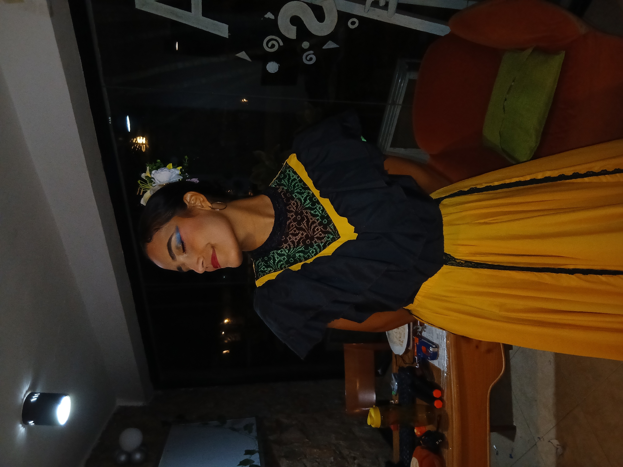 A woman in traditional dress is the focal point of the image. The dress is yellow and black with lace detailing around the waist. She wears a flower in her hair and makeup. The setting appears to be indoors, near a window with reflections of outdoor lights suggesting it's evening. There is furniture visible, including a chair and a table with various objects on it. The specific location, Playa El Agua, Venezuela, isn't apparent in the image.