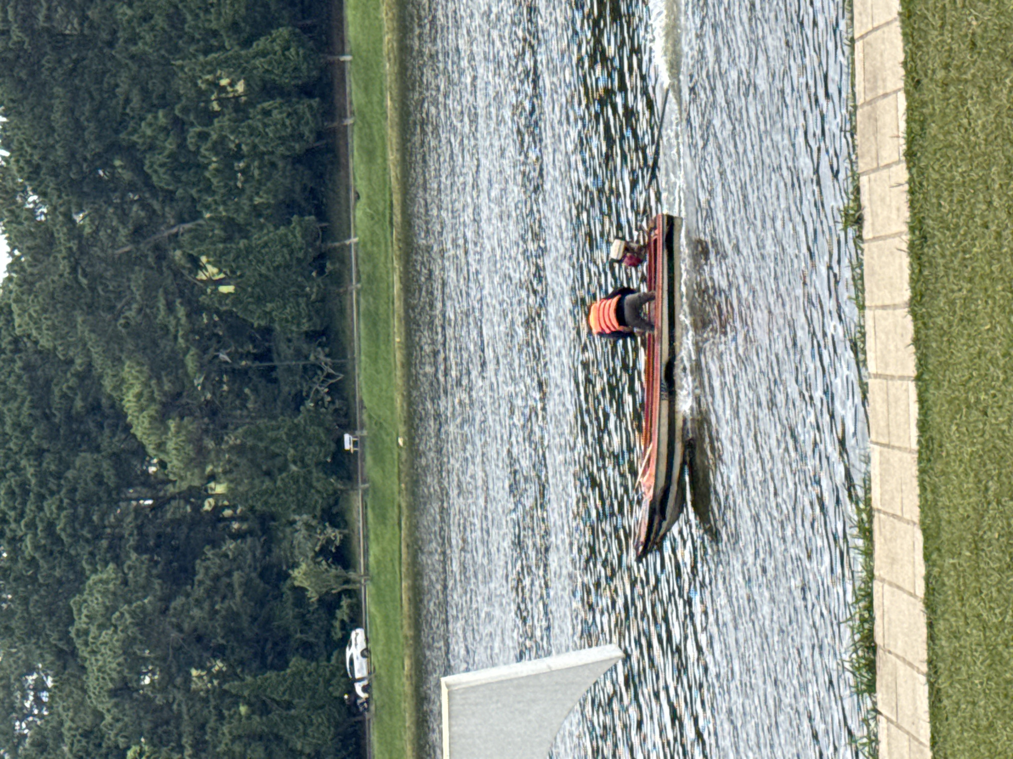 A man wearing an orange life vest and gray pants is operating a small motorboat on a lake in Da Lat, Vietnam. The boat, which has a red hull, is moving from left to right, leaving a wake behind it. The lake is surrounded by lush green trees, and in the background, there is a road with a white car. The foreground shows a grassy area with a paved walkway. The image captures a serene moment of activity on the water, with the natural beauty of Da Lat as the backdrop.