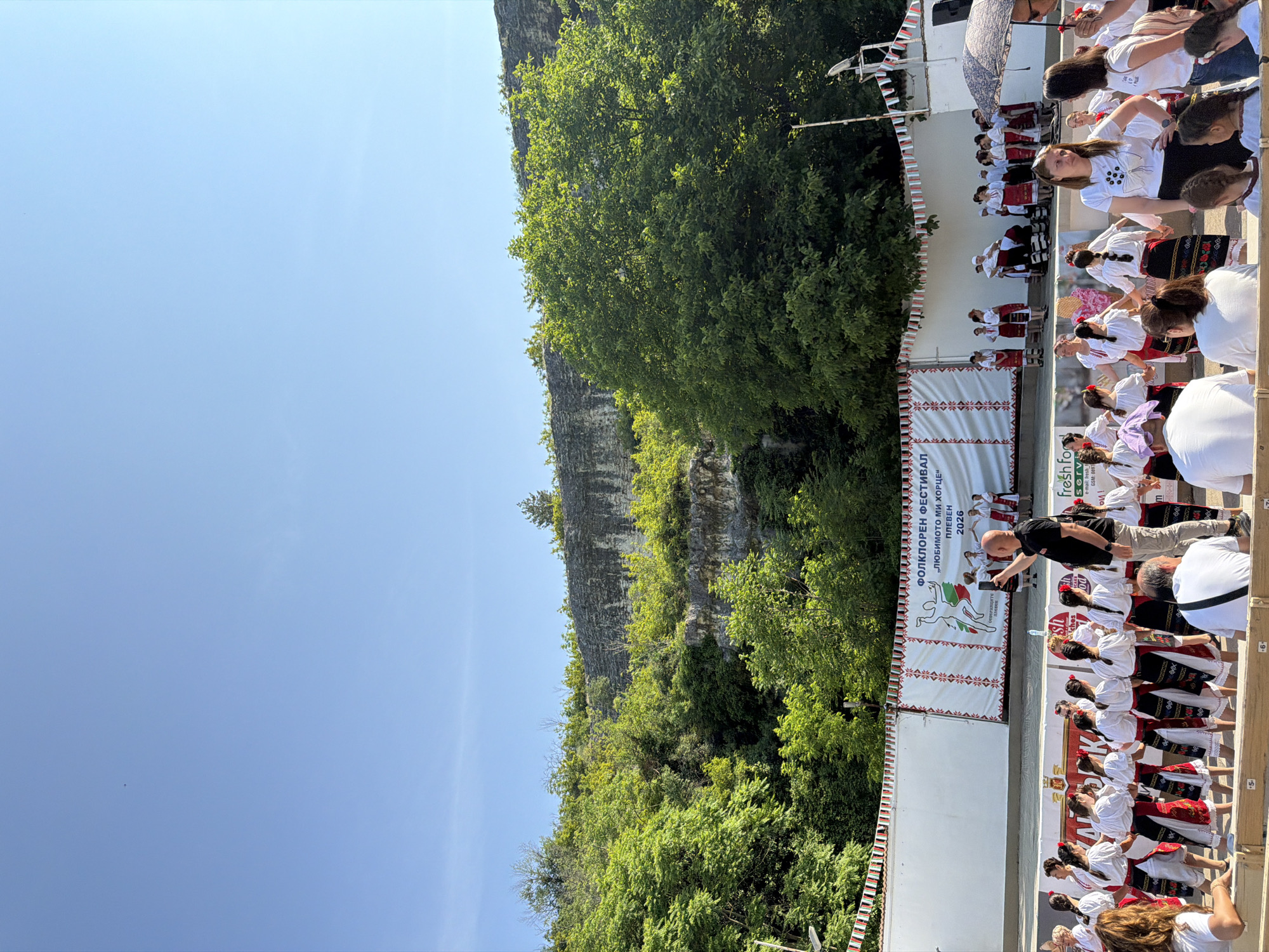 This image depicts an outdoor folklore festival in Pleven, Bulgaria. The scene is dominated by a clear, bright blue sky above a steep, rocky cliff face covered in dense green deciduous trees and shrubs, serving as a natural backdrop.
In the midground, a white stage is set up against the natural rock wall. A large banner on the stage reads "ФОЛКЛОРЕН ФЕСТИВАЛ „ЛЮБИМОТО МИ ХОРЦЕ“ ПЛЕВЕН 2026" (Folklore Festival "My Favorite Horo" Pleven 2026), flanked by vertical banners with traditional geometric patterns. A stylized dancer logo is visible on the main banner. Along the top of the stage and extending to the sides, red, white, and green bunting, reminiscent of the Bulgarian flag colors, is strung. On the stage, several groups of individuals, predominantly women and some men, are dressed in traditional Bulgarian folk costumes, consisting of white blouses, red and black patterned skirts or aprons, and often head coverings. They appear to be performing or preparing to perform a traditional dance. A speaker on a stand is visible to the right of the stage.
In the foreground, a large group of people is gathered. Many individuals, mostly women, are also dressed in traditional Bulgarian folk costumes, while others wear white t-shirts and dark bottoms. They are arranged in a large circular or semicircular formation, holding hands, indicative of a Horo dance. Some people are seated on wooden benches in the immediate foreground, observing the activities. One man in a black t-shirt and light-colored pants is standing prominently, holding a smartphone overhead, apparently recording or photographing the stage performance. To the far right, a person holds a large dark umbrella. A "fresh food" banner is partially visible in the lower right corner, suggesting a sponsor or vendor. Children are interspersed among the adults in the foreground. The ground appears to be a cleared, flat area suitable for dancing. Bright sunlight casts shadows, indicating a sunny day.