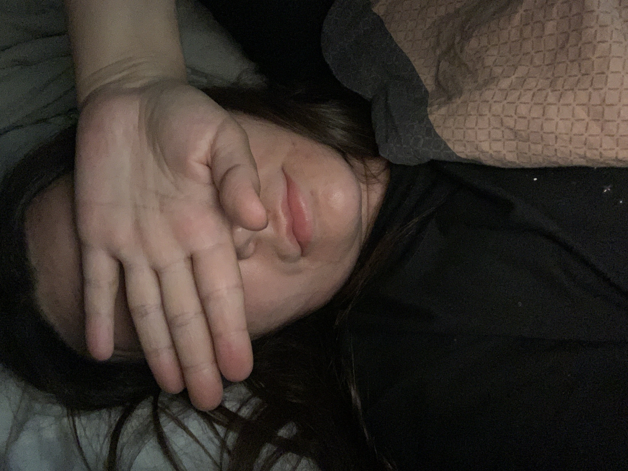 A close-up, overhead shot shows a person's face partially obscured by their hand. The person is lying on their side, with their face turned towards the viewer. Their eyes are closed, and their lips are slightly parted, as if they are sleeping or resting. The person's hand is positioned over their eyes, covering most of their face. The background is a dark, blurry surface, possibly a bed or a pillow. The lighting is dim, casting shadows on the person's face and hand.