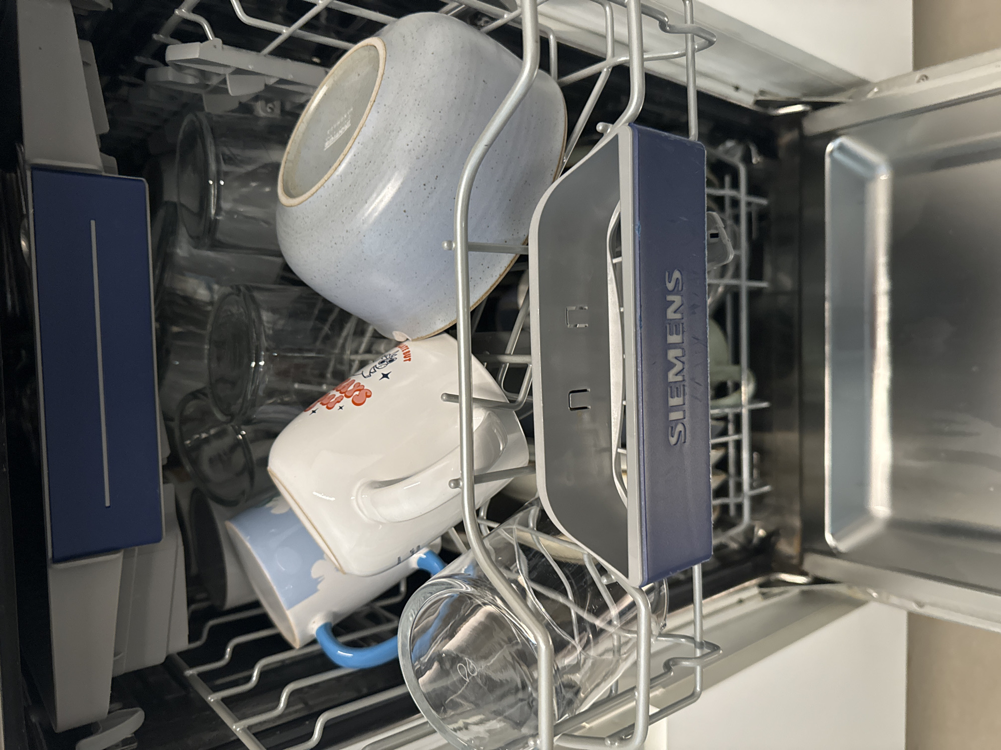 This media file shows the inside of a dishwasher filled with various items. The subjects are primarily objects, including several white ceramic mugs, one with red and blue floral designs and another with a blue cloud pattern, a large pale blue ceramic bowl, and clear glass tumblers. A blue rectangular object with the text "SIEMENS" is also visible. The setting is the interior of a dishwasher, with metal racks holding the dishes. The activity taking place is the loading of dishes into the dishwasher, presumably in preparation for a wash cycle. The lighting suggests it is daytime, and the overall impression is one of domestic organization. There are no people visible, and no specific indications of weather or emotions. The location cue is provided as Murcia, Spain.