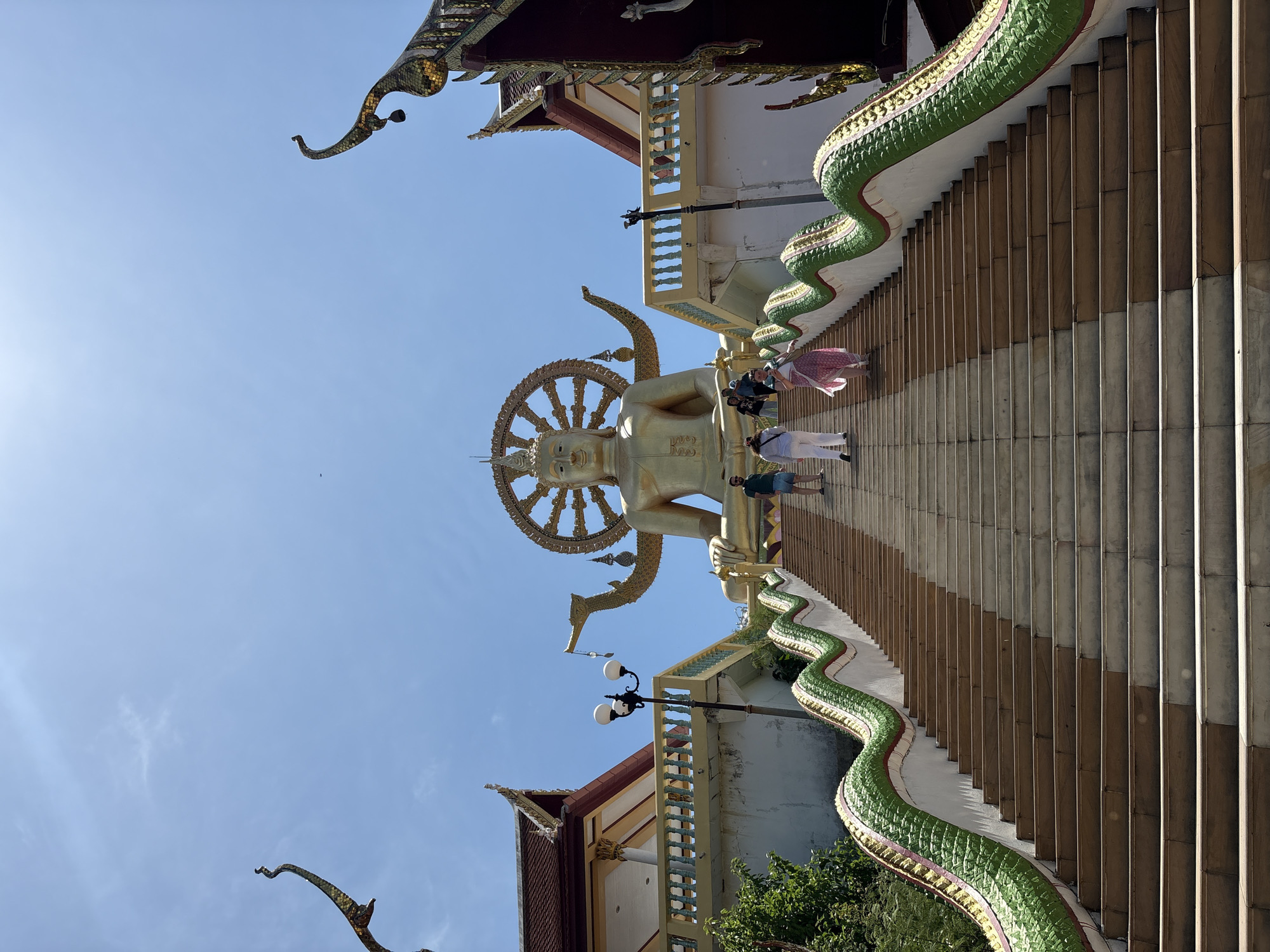 The image shows the Big Buddha statue at Wat Phra Yai in Baan Plai Laem, Thailand, viewed from below. A golden statue of Buddha stands prominently at the top of a staircase, adorned with a large, ornate halo or wheel behind its head. Several tourists are visible on the staircase. The staircase is flanked by green serpent sculptures. The sky is clear and blue. Traditional Thai architectural elements are visible on the surrounding buildings.
