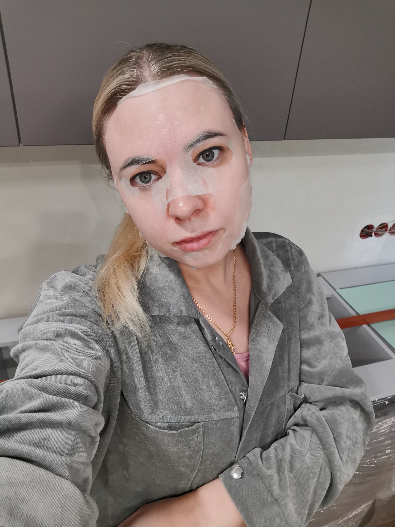A woman wearing a facial mask is taking a selfie in what appears to be an indoor environment. The location is potentially in the city of Chaikovsky, Russia. She has blonde hair pulled back in a low ponytail and is wearing a gray corduroy jacket and a gold necklace. A pink shirt peeks out from under the jacket. The background is comprised of what seems to be kitchen cabinets and a section of a counter or table. The photo is taken at a close range, with the woman's face being the primary focus. In the upper right corner, there's evidence of some electrical wiring or conduits visible in the wall.