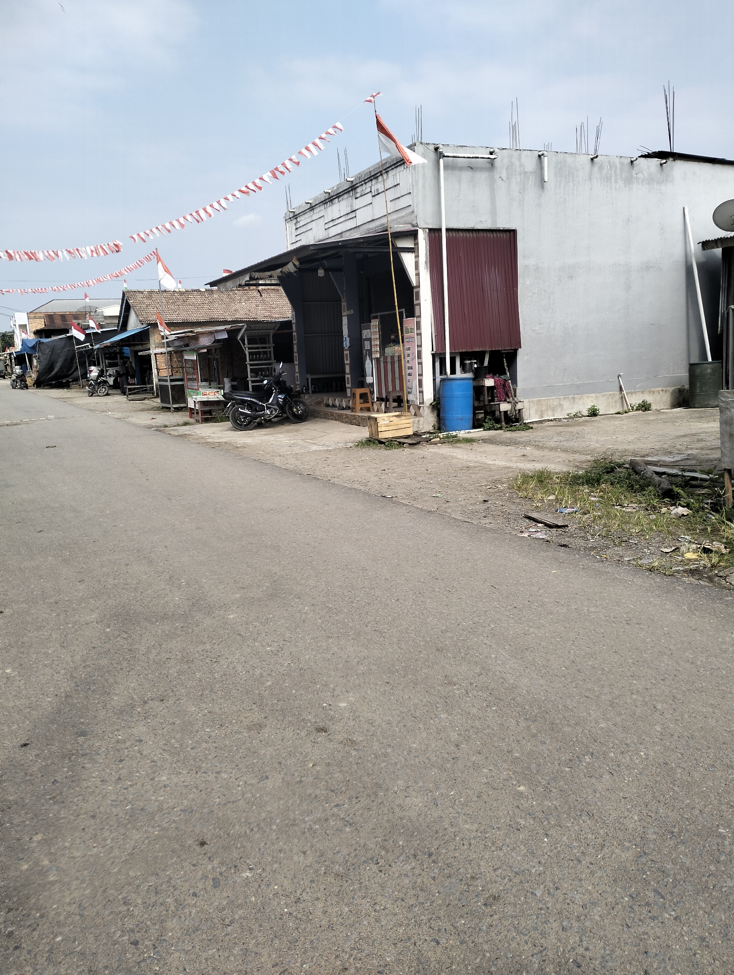 The image depicts a street scene in Pematang Lumut, Indonesia, likely decorated for a national celebration, given the numerous Indonesian flags and red and white pennant banners strung across the street and displayed on poles. The street itself is paved with asphalt and appears to be a two-lane road.
On the right side of the street, a light-colored, two-story building with exposed rebar at the roofline is visible. It has a dark red corrugated metal section on its facade and a large blue barrel next to it. Adjacent to this building are a row of small shops or stalls, characterized by their simple structures with awnings and open fronts. One stall displays a black motorcycle parked in front. Further down the row, more stalls are visible, some with blue tarps covering them. Several other motorcycles are parked along the side of the road.
The left side of the street is less developed, with grassy areas and scattered debris. The sky is partly cloudy. The overall impression is of a typical roadside commercial area in a smaller Indonesian town or village.