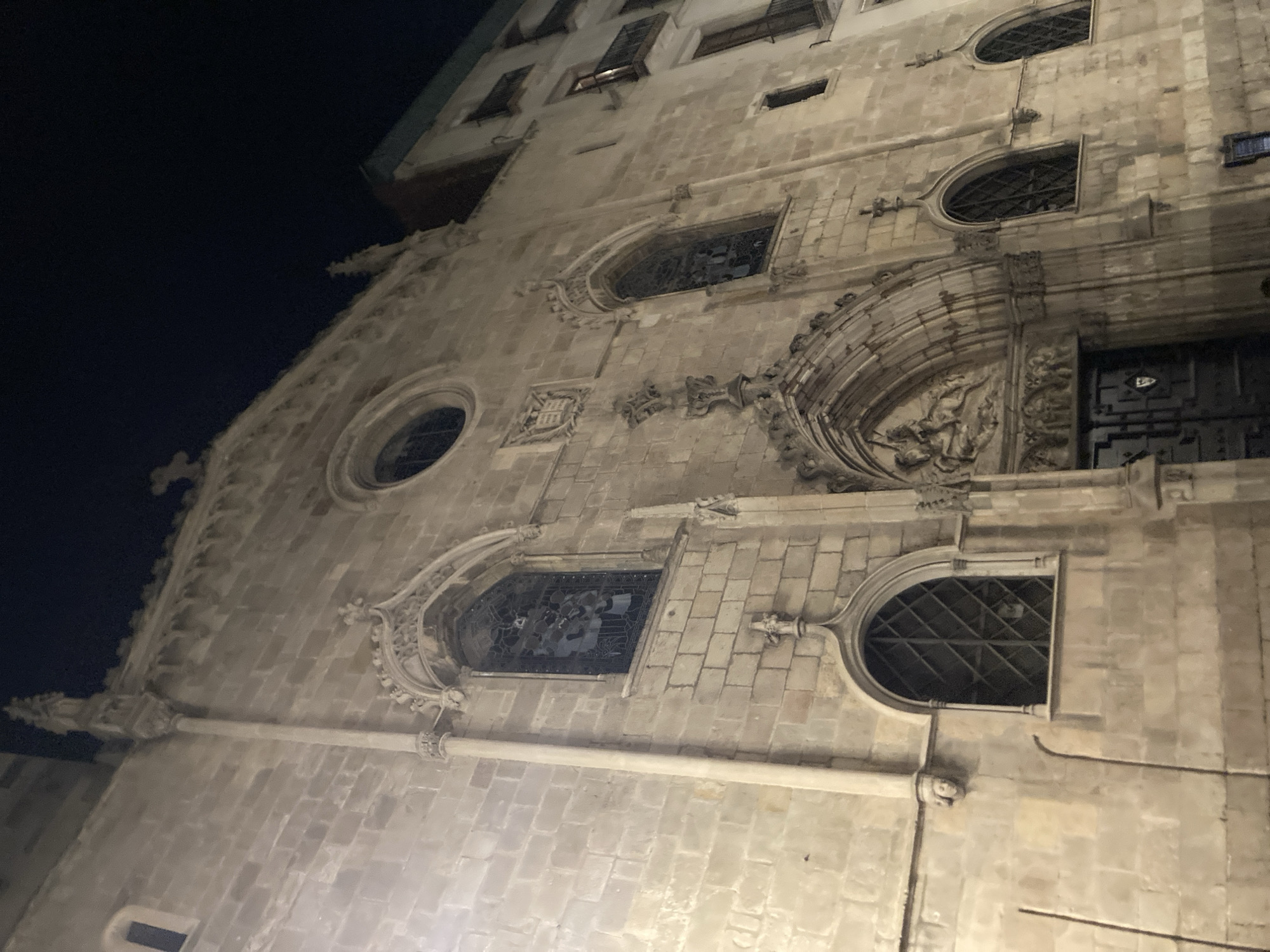 The image shows the facade of a stone building, likely a church or cathedral, at night. The architecture appears to be Gothic, with pointed arches, decorative tracery, and stained-glass windows visible. There is an ornate entrance with a sculptural relief above it. The building is illuminated by artificial light, casting shadows and highlighting the texture of the stone. In the background, parts of other buildings are visible, one of which has balconies. The sky is dark, indicating nighttime. The scene appears to be a public space in a city.