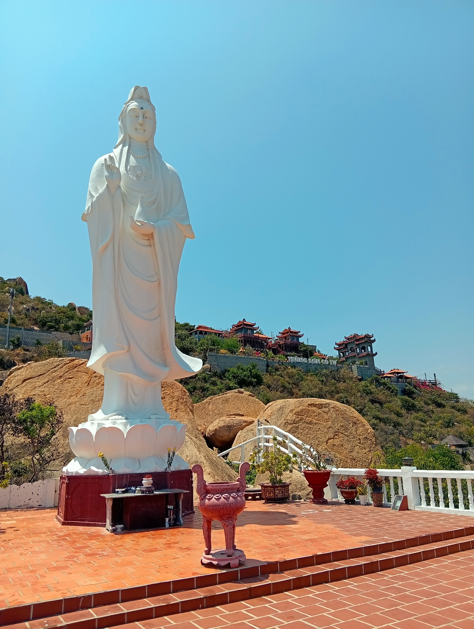 A tall, white statue of a standing Guanyin, a Buddhist deity, is the central focus of this image. The statue stands atop a red pedestal adorned with a lotus flower design, and a table with offerings rests in front of it. An ornate incense burner stands to the right of the pedestal.
The statue is situated on a brick-paved terrace with a white balustrade in the background. Behind the terrace, a hillside with large boulders is visible. Several traditional Vietnamese pagodas or temples with red roofs and tiered architecture are nestled on the hillside. The vegetation is sparse, consisting of low-lying trees and shrubs. The hillside has writing on it, "TUNG SON CO TU".
The sky is clear and bright blue, indicating a sunny day. The statue casts a shadow on the terrace, further suggesting direct sunlight. The overall setting appears to be a religious site or temple complex in Khánh Hải, Vietnam.