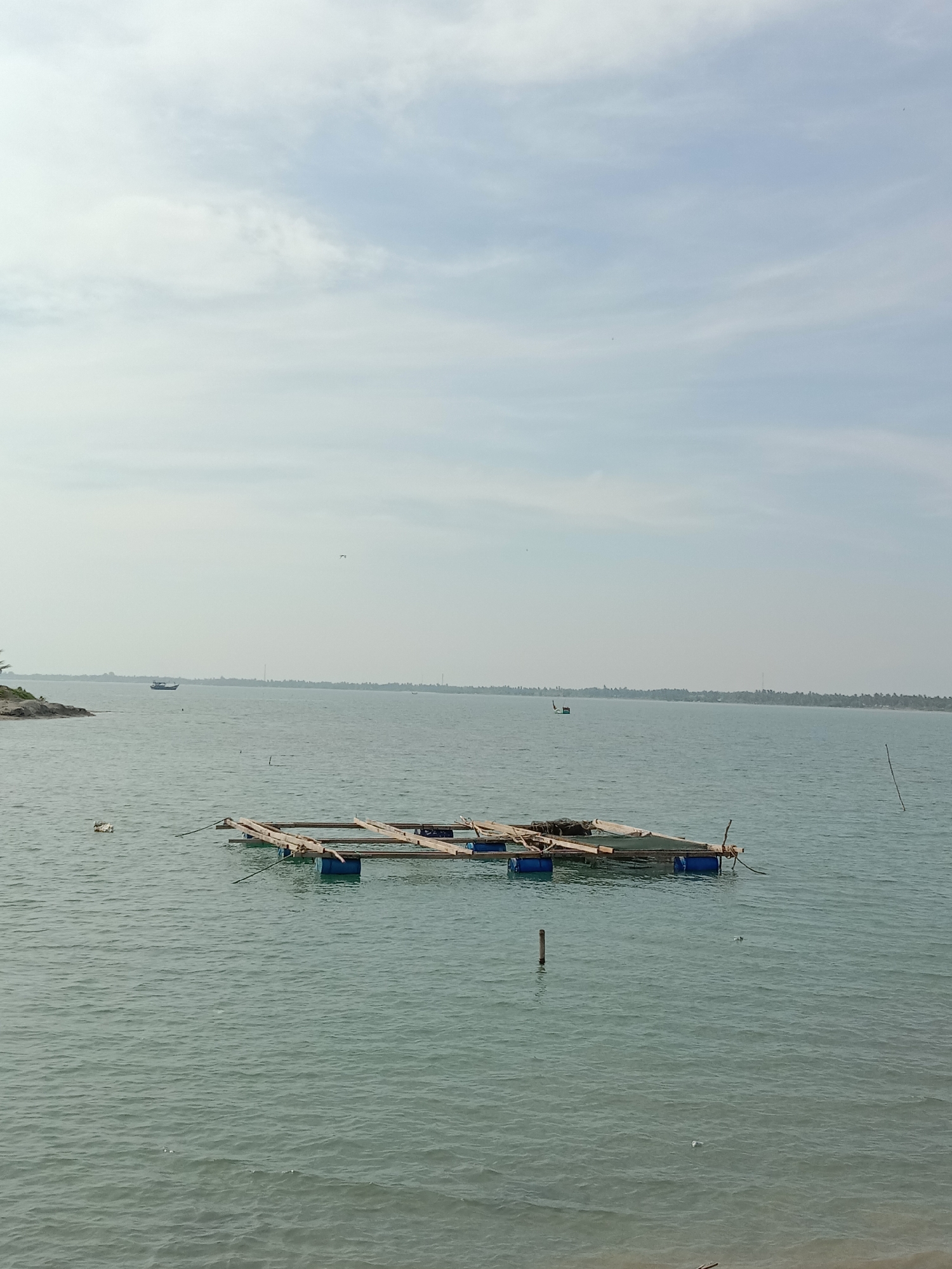 The image captures a serene, expansive body of water under a bright, overcast sky. The horizon features a distant, low-lying landmass lined with dense trees, likely palms, stretching across the entire background.
In the foreground, slightly to the left of the center, a floating structure made of wooden beams and blue barrels is visible, possibly serving as part of a fish farm or a simple raft. Several thin wooden poles protrude from the water at various points, and some light-colored debris, possibly plastic, floats near the raft and further out.
In the mid-ground, a dark blue boat is seen navigating the calm waters towards the left, with another less distinct boat visible further in the distance. A small bird is captured in flight in the upper central part of the sky. The water's surface has gentle ripples, reflecting the soft, diffused light from the cloudy sky.
The time of day appears to be daytime, likely late morning or early afternoon, given the overall brightness without harsh shadows. The weather is overcast or partly cloudy, creating an even, diffused light across the scene. The tranquil water and distant land suggest a coastal or estuarine environment. No people are visible, and there is no discernible text in the image. The scene conveys a peaceful atmosphere, characteristic of a working waterfront in Lhokseumawe, Indonesia.