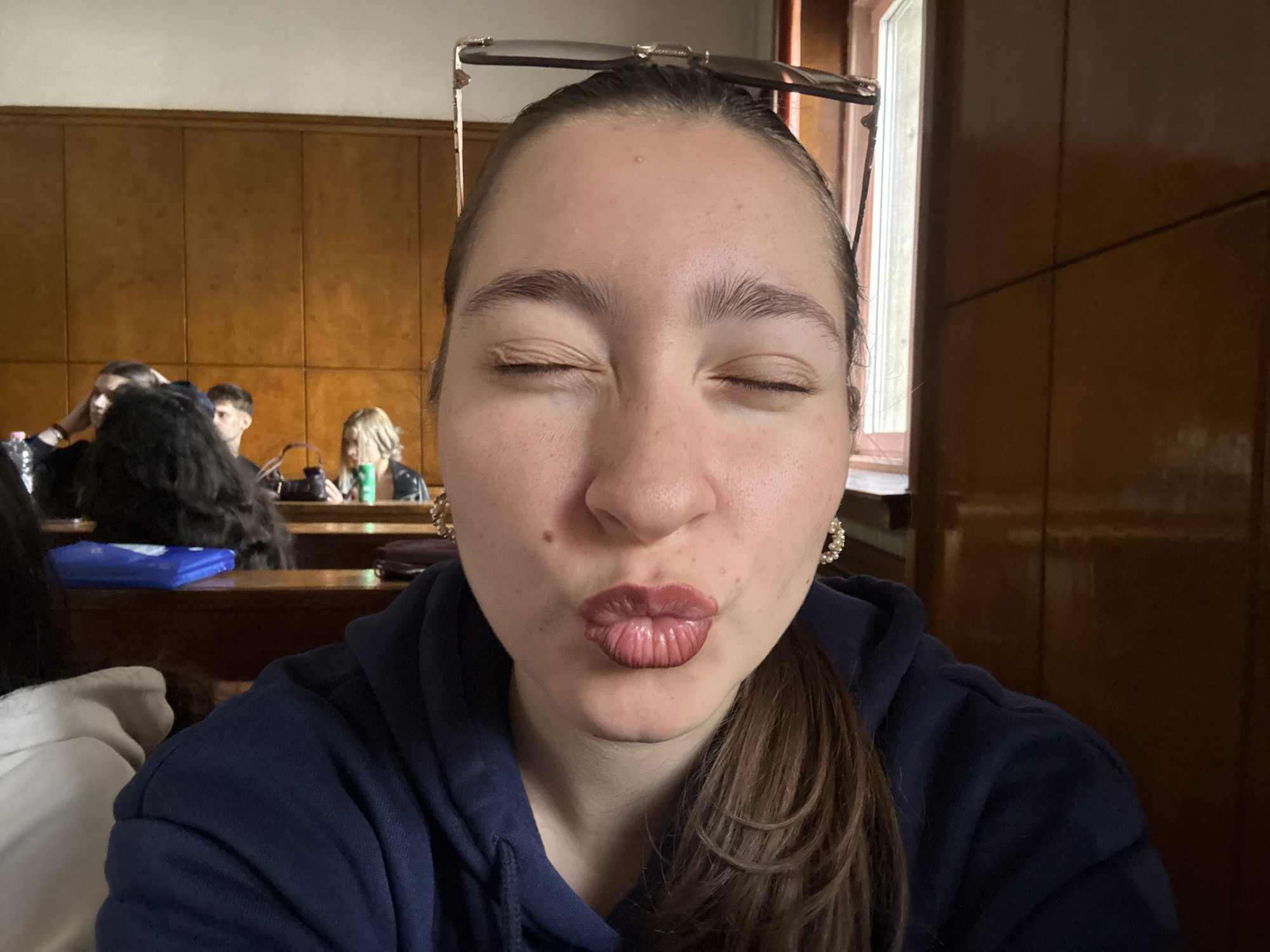 A fair-skinned woman with brown hair pulled back into a ponytail, wearing a dark blue hooded sweatshirt and small pearl earrings, is posing with her eyes closed and lips pursed in a “kiss” expression. Sunglasses rest on her forehead. She is in a classroom or lecture hall with wood paneling behind her. Other students are seated in rows behind her. The location is likely a university classroom in Sofia, Bulgaria.