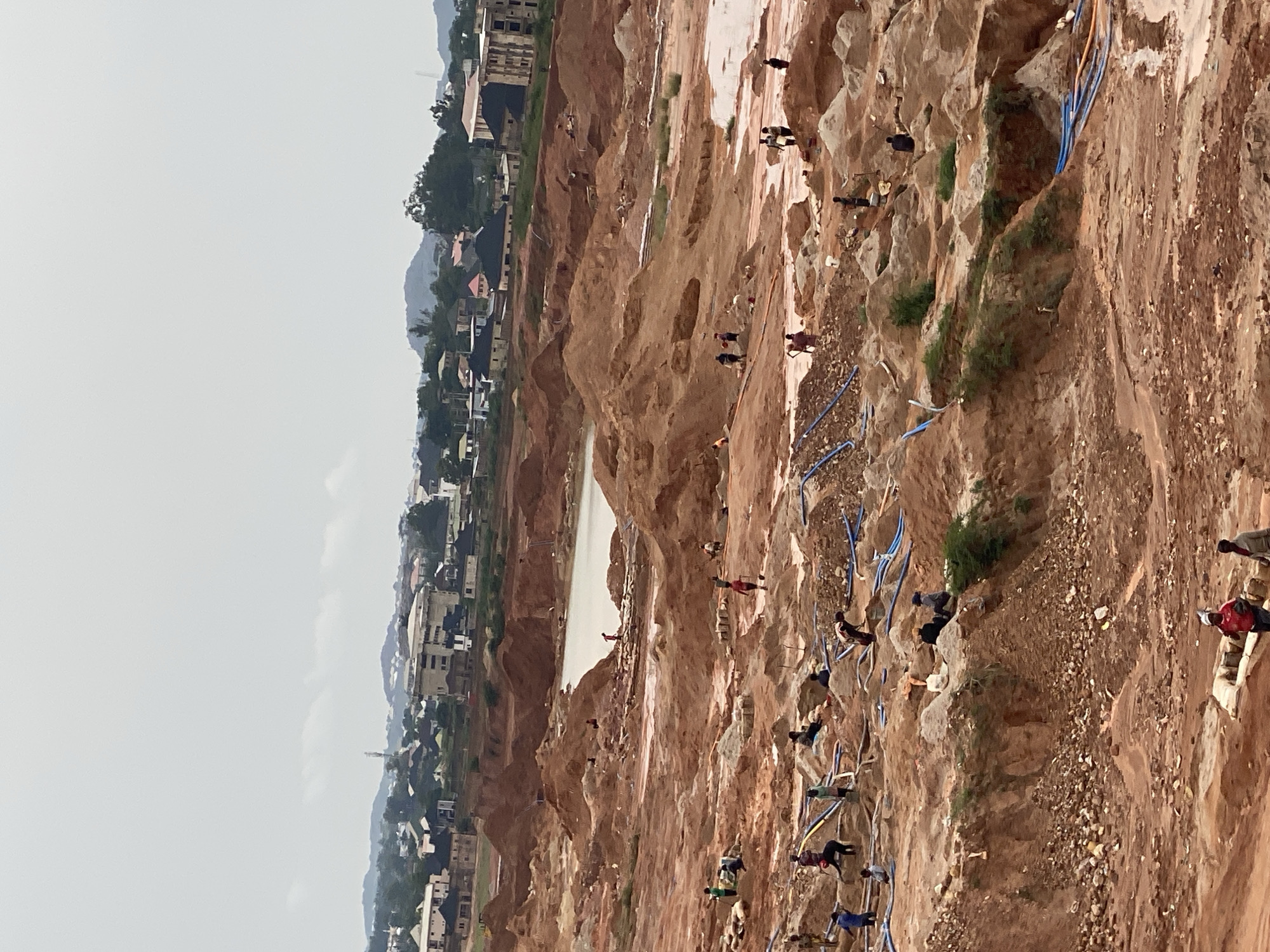 This image depicts a vast, excavated area resembling a mining or construction site, likely located in or near the city of Jos, Nigeria. The terrain is characterized by large mounds of reddish-brown soil and sand, with evidence of recent digging and earthmoving. Several individuals, appearing to be laborers, are scattered across the site, engaged in various activities. Some are seen carrying objects, digging, or standing amidst the earth. Blue pipes are visible laid out on the ground, possibly for water distribution or drainage. In the background, a body of water, likely a pond or pool formed by the excavation, is present. Beyond the immediate site, a developing urban landscape with buildings and hills can be observed, suggesting the proximity of a residential or commercial area. The sky is overcast, contributing to a subdued lighting condition across the scene.