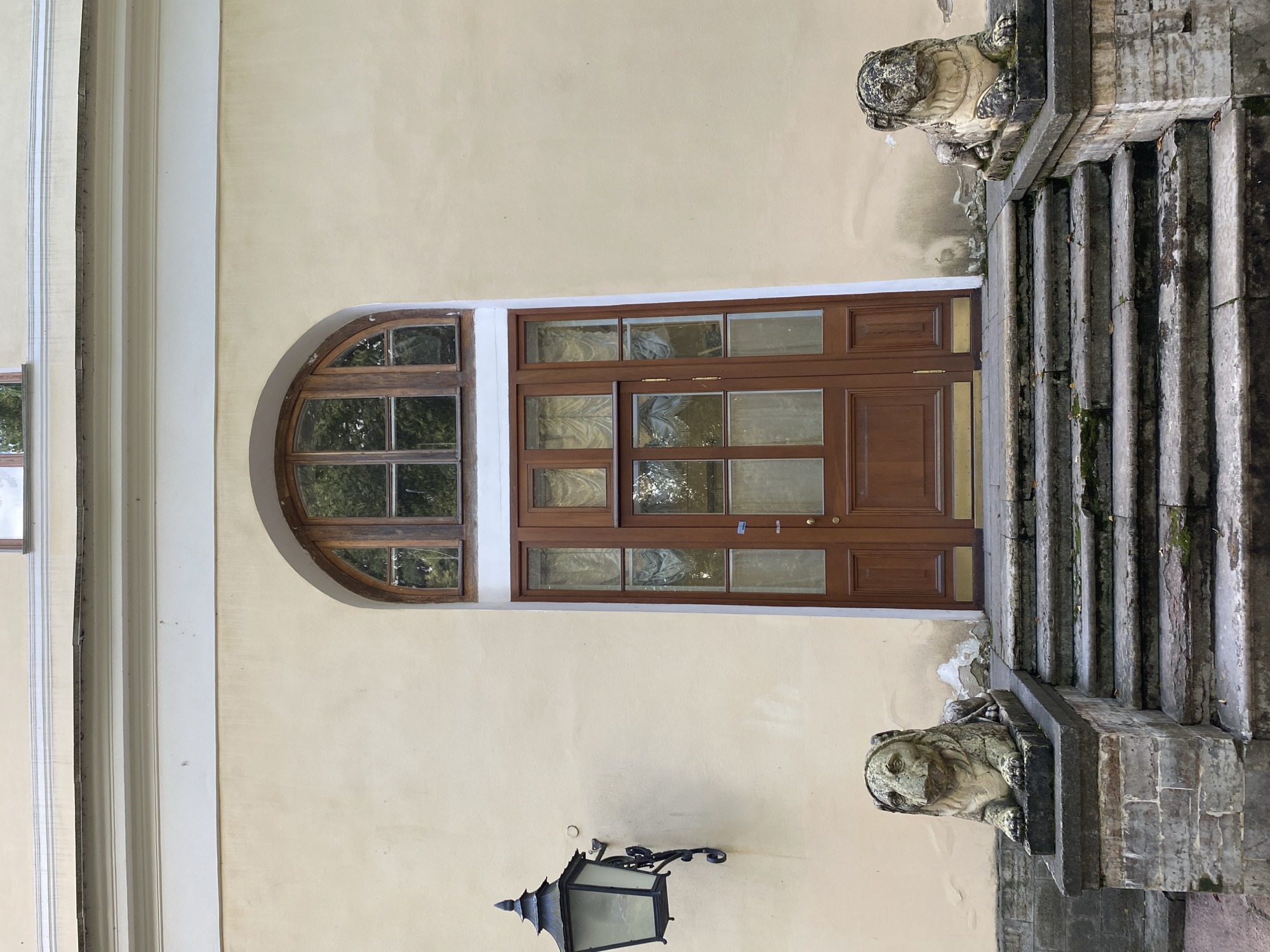 The image shows the facade of a building with a large arched window above a wooden double door. The building has a cream-colored stucco wall. To the left of the door, an ornate black metal lantern is mounted on the wall. In front of the door, a stone staircase leads up to the entrance. On either side of the staircase, there are stone sculptures of lions. The lion on the left is partially visible. The lion on the right is fully visible, seated on a stone pedestal. The staircase and pedestals are made of rough-hewn stone blocks, showing signs of age and weathering with moss and debris. The setting appears to be a historical park or estate. This is the entrance to the Chinese Palace in Pavlovsk, Russia.