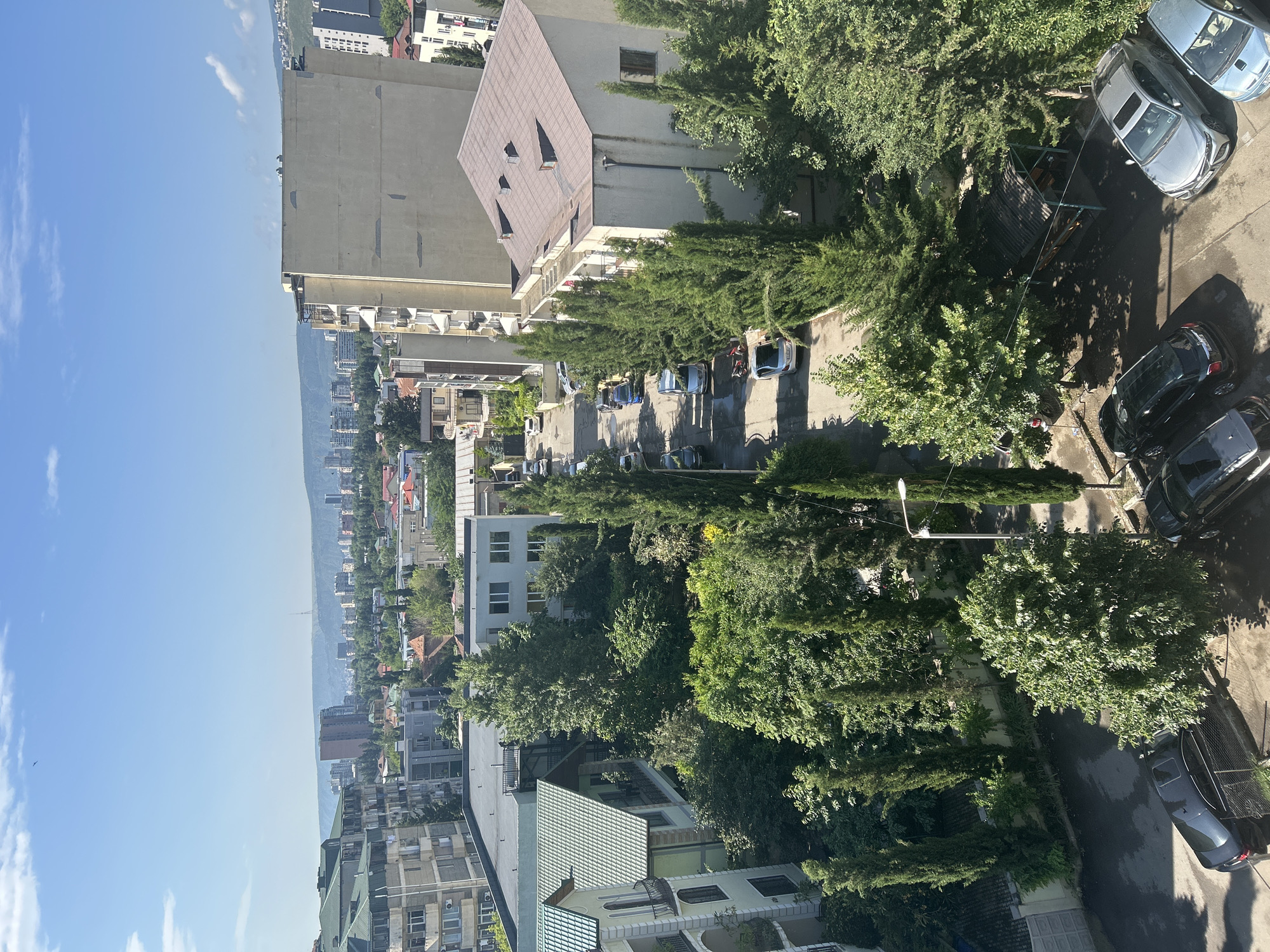 The image is an outdoor, elevated shot of a residential street in a city on a sunny day. The street is lined with various buildings, including apartment blocks and houses, interspersed with abundant green trees and cypress trees. Several cars are parked along the street and in a parking area. A small, covered seating area with tables is visible in the lower right portion of the image. The sky is blue with a few scattered white clouds. In the distance, a hazy cityscape and rolling hills are visible under the bright sun.