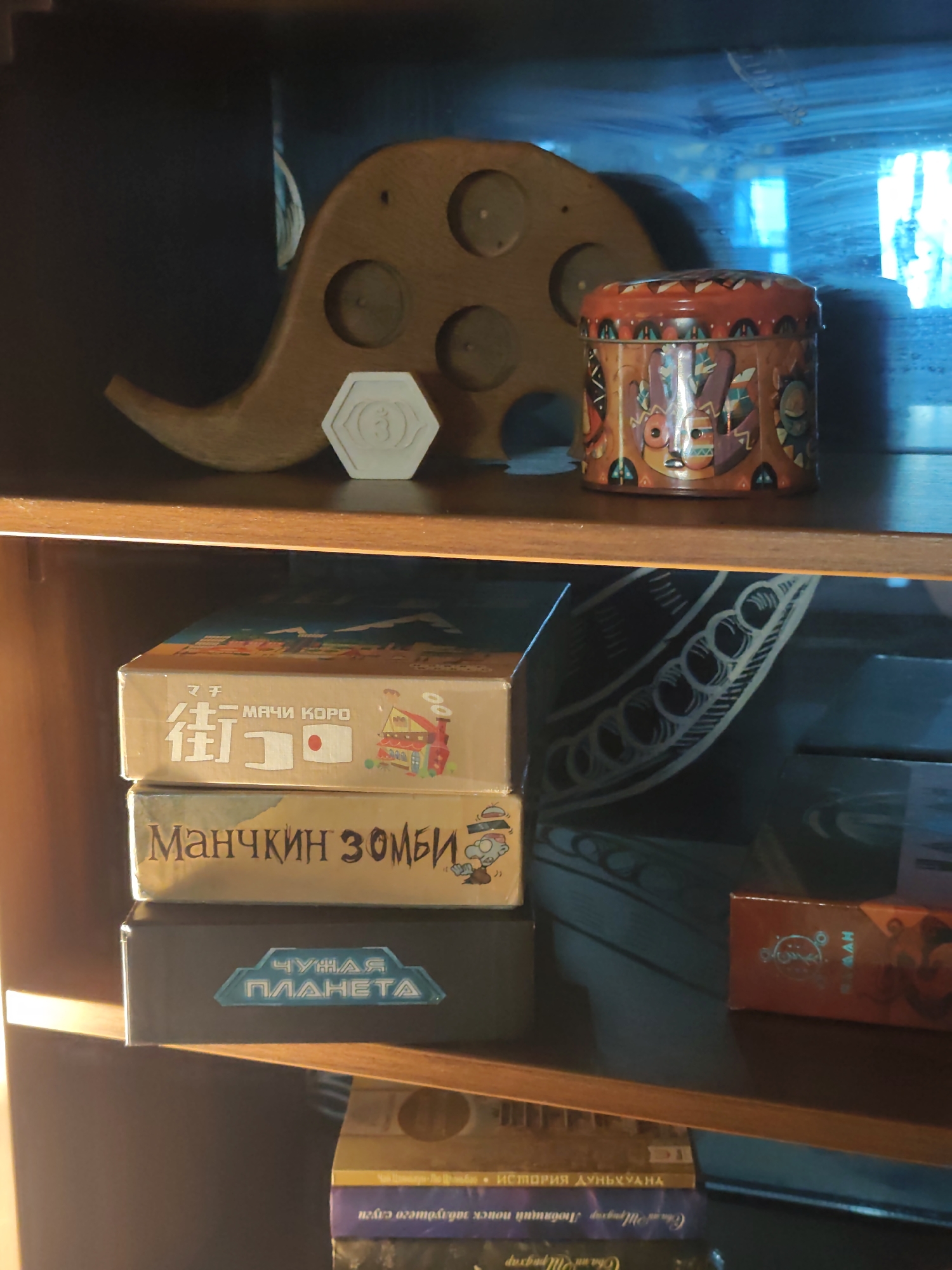 A medium shot captures a collection of objects on a wooden shelf, illuminated by warm, side lighting. The upper shelf displays a carved wooden elephant-shaped object with circular indentations, a hexagonal white token with a circular symbol, and a cylindrical tin box decorated with colorful, abstract patterns. The lower shelf is occupied by three stacked game boxes. The top box features Japanese characters, the word "Machi Koro" in English, and a cartoon illustration of a house and train. The middle box is labeled "Manchkin Zombies" in Russian. The bottom box is dark gray with a light blue hexagonal emblem containing the Russian words "Chuzhaya Planeta". Further down, partially obscured, are more book-like objects, one with visible Russian text. The background is a dark, reflective surface with abstract, circular patterns. The overall scene suggests an indoor setting, possibly a home or shop, with the warm lighting creating a cozy atmosphere. There is no visible indication of the city of Sochi, Russia.