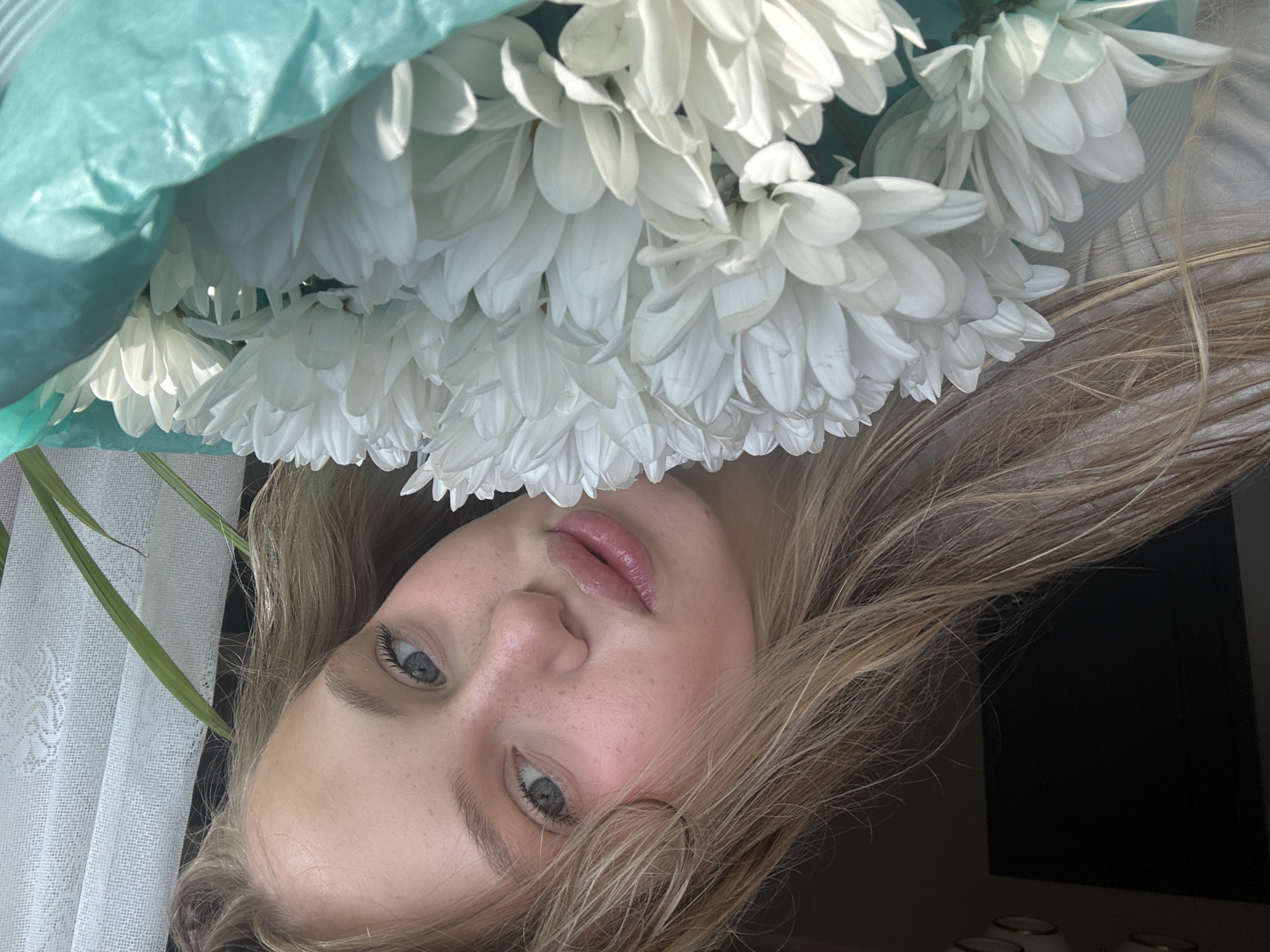 A young blonde woman with fair skin and freckles is looking at the camera, holding a bouquet of white chrysanthemums. Her eyes are blue, and she is wearing light-colored clothing. She is positioned in the foreground, with the flowers obscuring the lower part of her face. In the background, there is a dark area, possibly a doorway, and a sheer white curtain with a lace pattern. A green plastic wrapping is visible around the base of the flowers. The lighting suggests an indoor setting with natural light coming from the side.