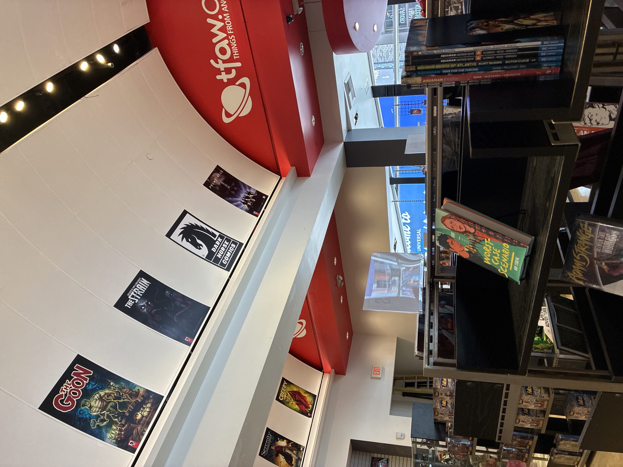 The image shows the interior of a comic book store. Various comic book covers and posters are displayed on the white, arched ceiling. In the foreground, there are bookshelves filled with comic books and graphic novels. One book prominently displayed on a shelf in the foreground is titled "WORST CASE SCENARIO" by Ray Stieve. To the right, a stack of comic books with various titles, including "AQUAMAN," are visible. A red sign with a white planet logo and "tfaw.com THINGS FROM ANOTHER WORLD" is mounted on a curved red structure above the bookshelves. An "EXIT" sign is visible on a light-colored wall in the background, to the left of a large screen displaying a blue graphic. The store appears to be well-lit with artificial lighting.