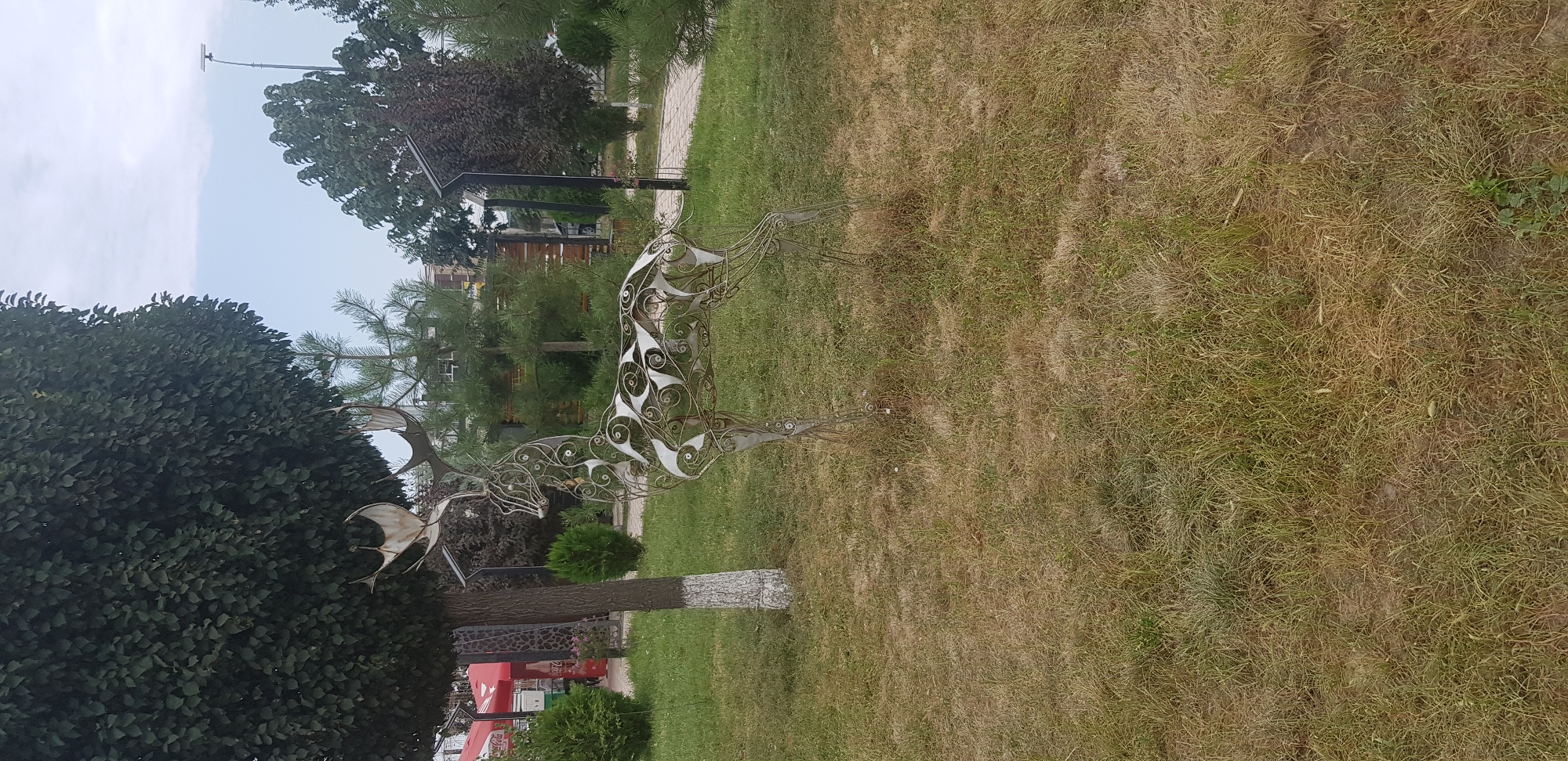 An outdoor, medium shot captures a wire sculpture of a deer standing on a patch of dry grass in front of a large, leafy tree. The deer sculpture is intricate, made of thin metal wires forming its body, legs, and antlers, with decorative swirling patterns on its sides. The tree is tall and dark green, with its trunk partially painted white. Behind the deer and tree, a path is visible, with more trees and greenery, including some dark red-leaved bushes, and buildings in the distance. A street lamp is also visible in the background. The sky is overcast.