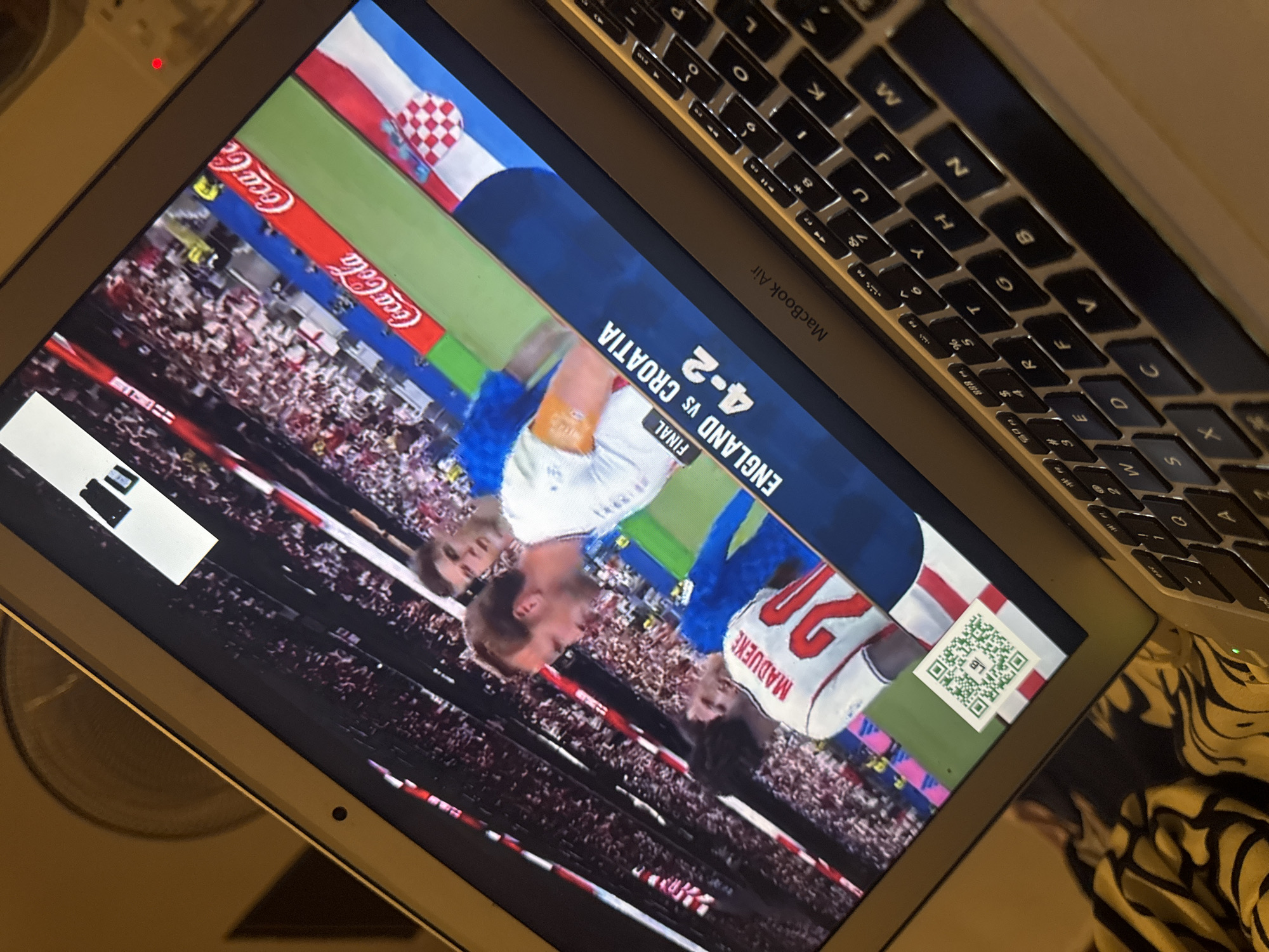An overhead, angled shot captures a laptop screen displaying a soccer match, likely a recording or live stream. The screen shows players in white and blue jerseys, with one player in white bearing the name "MADUENE" and the number "20" on his back. A graphic on the screen indicates "FINAL ENGLAND VS CROATIA 4-2," along with Coca-Cola advertisements along the sidelines and the Croatian flag. A QR code is visible in the bottom left corner of the screen. The laptop's keyboard, with black keys and silver accents, is visible in the foreground. The background is blurred, suggesting an indoor setting. There is no discernible information to place this image in Kufena, Nigeria.