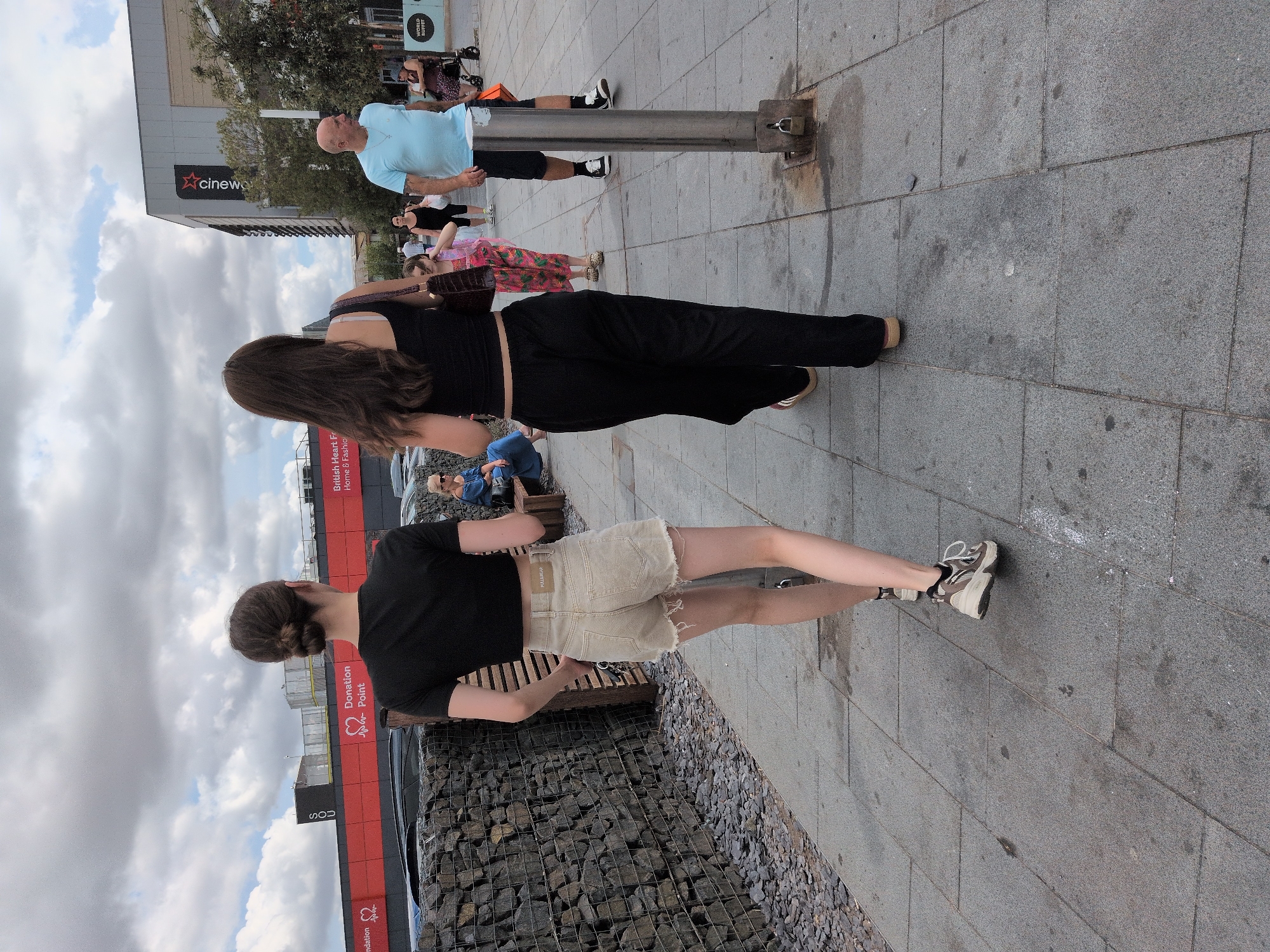 Two young women walk away from the camera on a paved sidewalk in what appears to be an urban area, possibly a shopping precinct. The woman on the left wears a black crop top and light-colored denim shorts, with her hair in a bun. The woman on the right wears a black tank top and black trousers, with long dark hair.
In the background, there is a building with a red and black facade featuring the text "British Heart Foundation Donation Point Home & Fashion." Above this, a cloudy sky is visible. Further to the right, a modern building with the "Cineworld" logo is present. Other individuals are scattered in the background, including a man in a light blue t-shirt and dark shorts, a woman in a wheelchair, and a child in a brightly colored outfit.
The sidewalk is made of grey paving stones. To the left of the women, there is a textured wall constructed of wire cages filled with stones. A metal bollard with a padlock is positioned on the sidewalk between the two women. The overall scene suggests a daytime setting in a public outdoor space.
