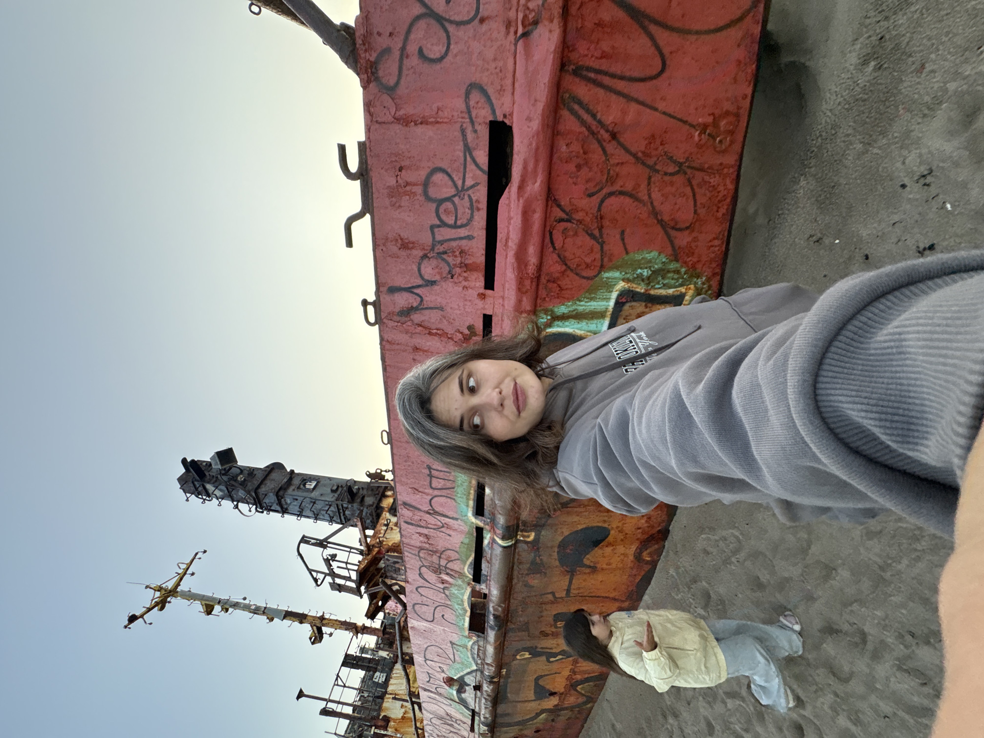 Two females, an adult and a child, are on a sandy beach in Teriberka, Russia, in front of a large, rusted shipwreck covered in graffiti. The adult, in the foreground, is taking a selfie with a gray hoodie. The child, in a yellow jacket and blue pants, stands to the left, gesturing with her right hand. The shipwreck dominates the background, its red hull and rusty metal structures weathered by the elements. The sky above is a pale blue.