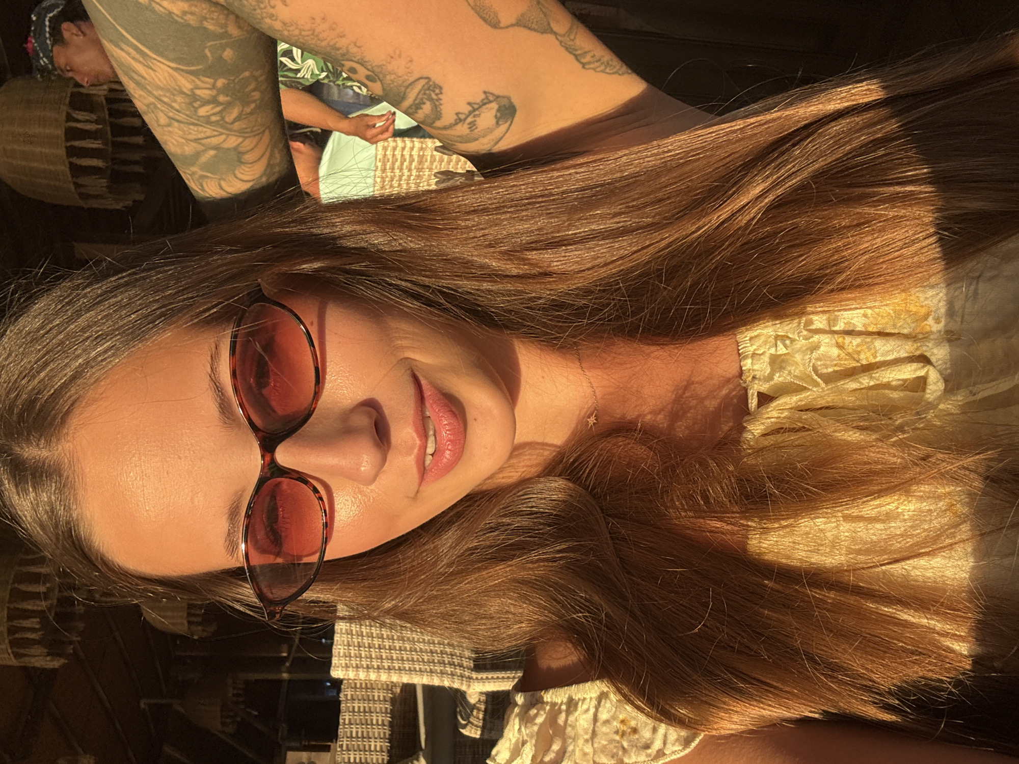 A medium shot depicts a fair-skinned female wearing red-tinted sunglasses. She has light brown hair and is smiling slightly. She is wearing a light-colored patterned top. In the background, a man with tattoos on his arm is visible. The location is North Kuta, Indonesia. The lighting suggests it is daytime. The picture appears to be taken indoors, possibly in a cafe or restaurant setting.