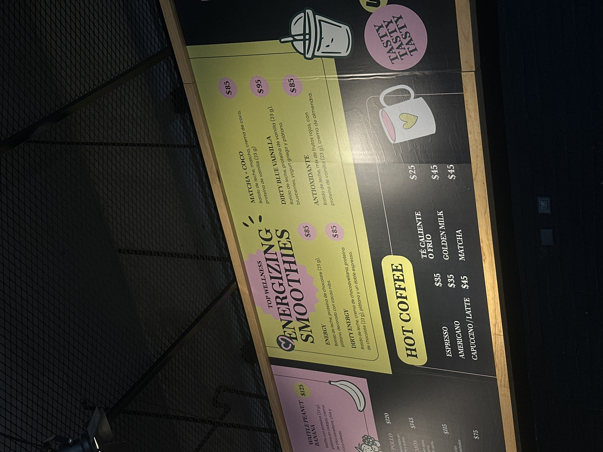The image shows a menu board with details about smoothies and hot coffee. The upper section highlights "Energizing Smoothies" with descriptions and prices for flavors like Matcha + Coco, Dirty Blue Vainilla, Energy, Antioxidante, and Dirty Energy. Below, the "Hot Coffee" section lists Espresso, Americano, and Capuccino/Latte, along with prices for hot or cold options, as well as Golden Milk and Matcha. The board includes a "Top Wellness" heading and features illustrations for the items. In the upper right corner, there is a "Tasty" graphic.
I am unable to provide information about the location of the image in the city of San Bernardino Tlaxcalancingo, Mexico.