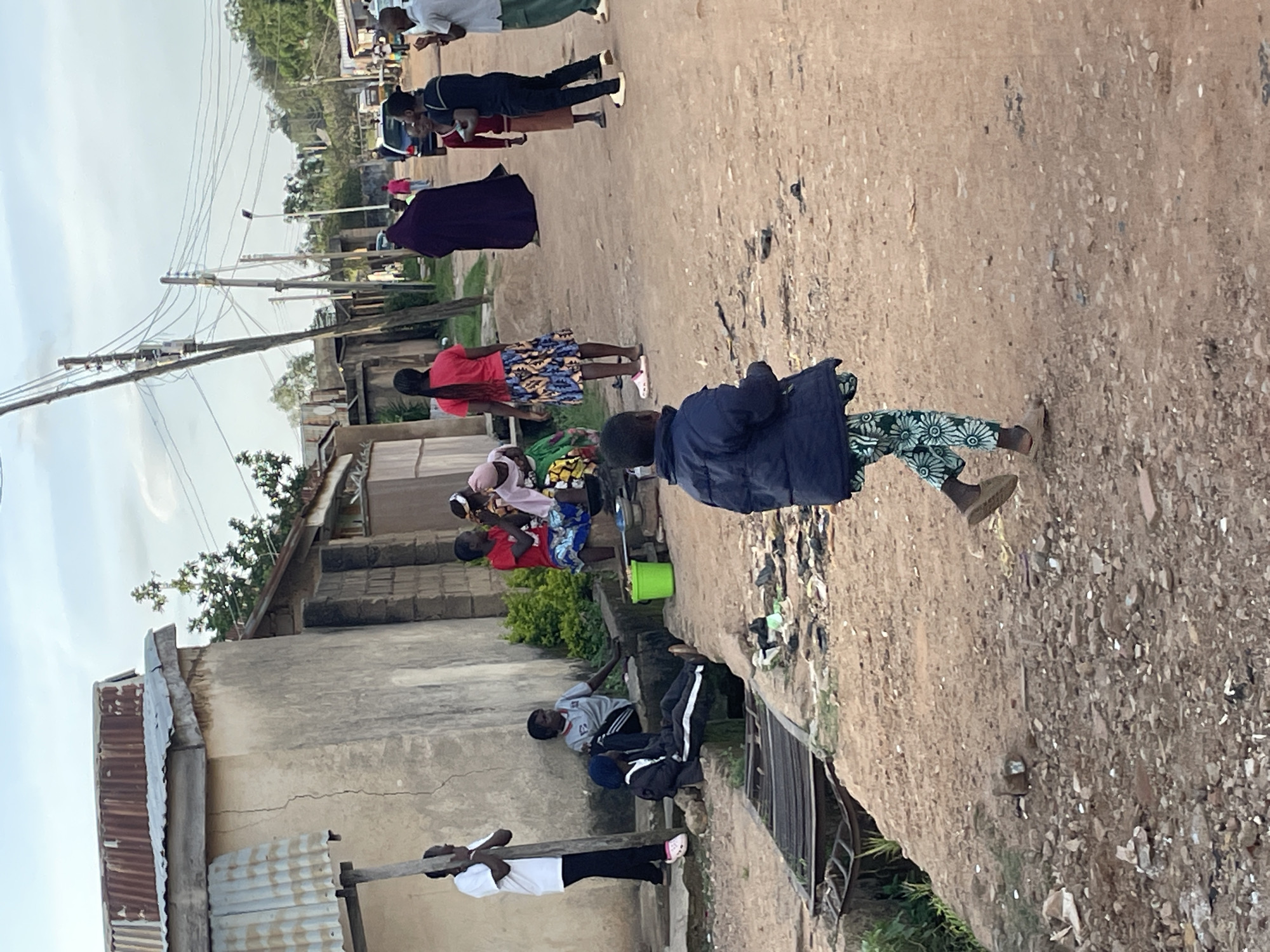 In Barakin Akawo, Nigeria, a street scene unfolds with a group of individuals gathered near a rough-hewn concrete building with a corrugated metal roof. A young boy in a dark blue jacket and patterned pants walks away from the camera on a dirt road strewn with debris and small stones. To his left, a woman in a vibrant patterned skirt and red shirt stands with her back to the viewer, facing a group of people sitting or standing near a drainage ditch. Among them, a person in a gray shirt with black stripes is seated, and another in a red shirt is visible. A green bucket rests near the ditch. Further down the street, several other individuals are dispersed, with a dark-colored car and more buildings visible in the background, along with telephone poles and a tangle of wires, suggesting an urban or semi-urban environment. The sky is overcast, with hints of diffused sunlight.