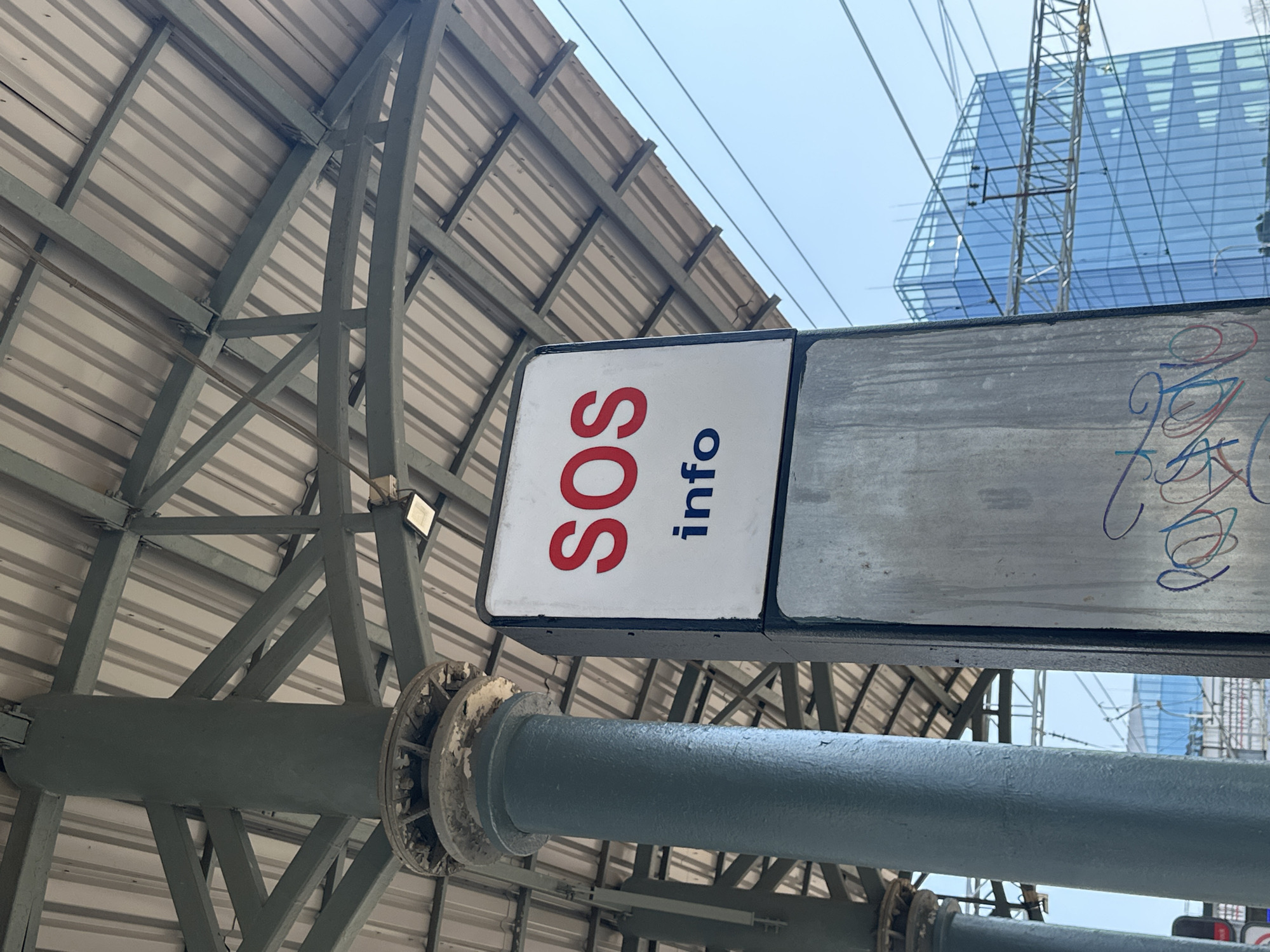 The image shows an SOS info sign on a metal pole in an outdoor urban environment, likely a train station. The sign has "SOS" in red letters and "info" in blue letters. The pole is grey and shows signs of wear and graffiti. Behind the pole, a large modern glass building is visible, suggesting a city location. Overhead, there is a metal roof structure supported by beams, characteristic of an outdoor public space like a station platform. Power lines are also visible in the background. The sky is clear and blue. There are no people visible in the frame. The overall scene is functional and utilitarian, with elements of both infrastructure and urban development.