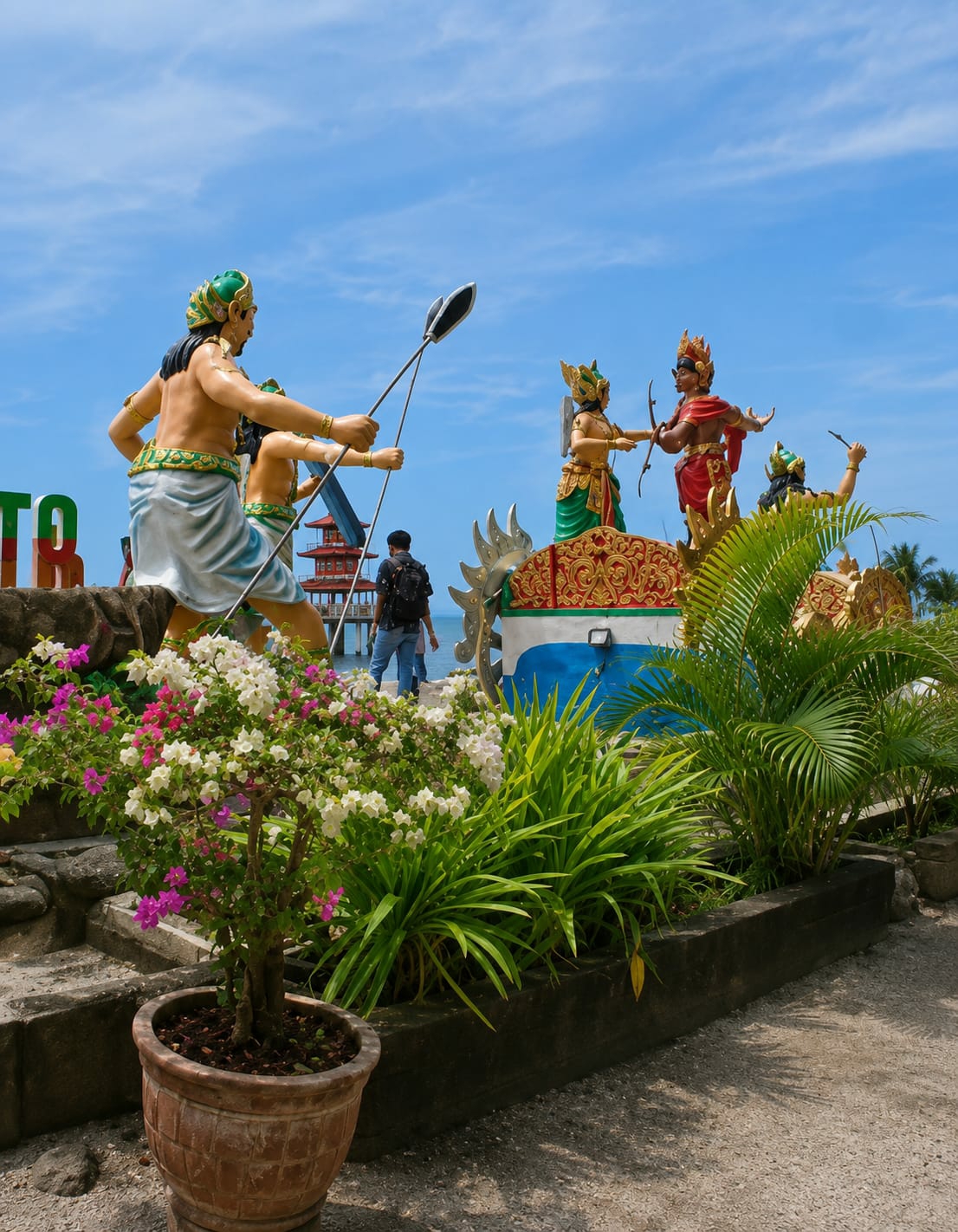 The image captures a scenic outdoor view at the Kota Pari attraction in Indonesia, under a bright blue sky with scattered clouds. Dominating the foreground is a vibrant bougainvillea plant in a terracotta pot, adorned with pink and white blossoms, nestled amongst lush green foliage.
In the background, a series of colorful statues depicting figures from what appears to be a mythological or historical scene are prominently displayed. One statue, a warrior in a green headdress and blue skirt, holds two spears, aiming them towards the right. To the right, two figures in ornate attire engage in what looks like a ceremonial gesture or perhaps a duel, one holding a bow and arrow. Another figure is visible further to the right, raising an arm.
A person wearing a black backpack and blue jeans walks away from the camera, past the statues, towards the sea visible in the distance. Signs with large green letters, partially obscured, are also present to the left, suggesting the name of the location. The overall setting is well-maintained, with stone pathways and decorative planters contributing to the attractive presentation of the Kota Pari attraction.