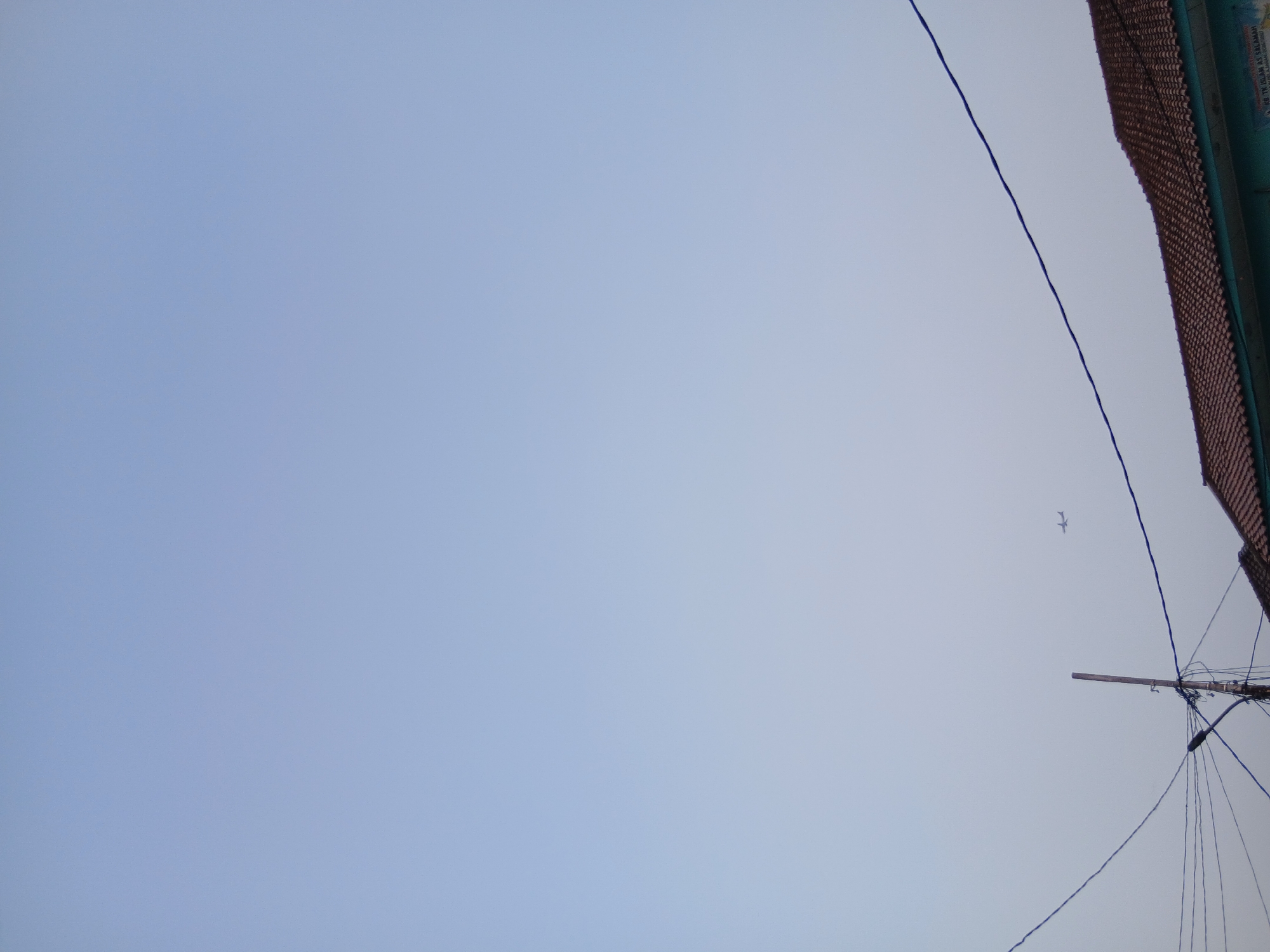 The image shows a pale blue sky with a small airplane in the distance. In the foreground, there are power lines strung between utility poles, a street lamp, and the tiled roof of a building. The building appears to be a school, indicated by faint text on a blue sign that reads "KB. TK ISLAM AS SALAMAH". The sky is hazy, suggesting either dawn, dusk, or atmospheric conditions. This scene is located in the city of South Tangerang, Indonesia.