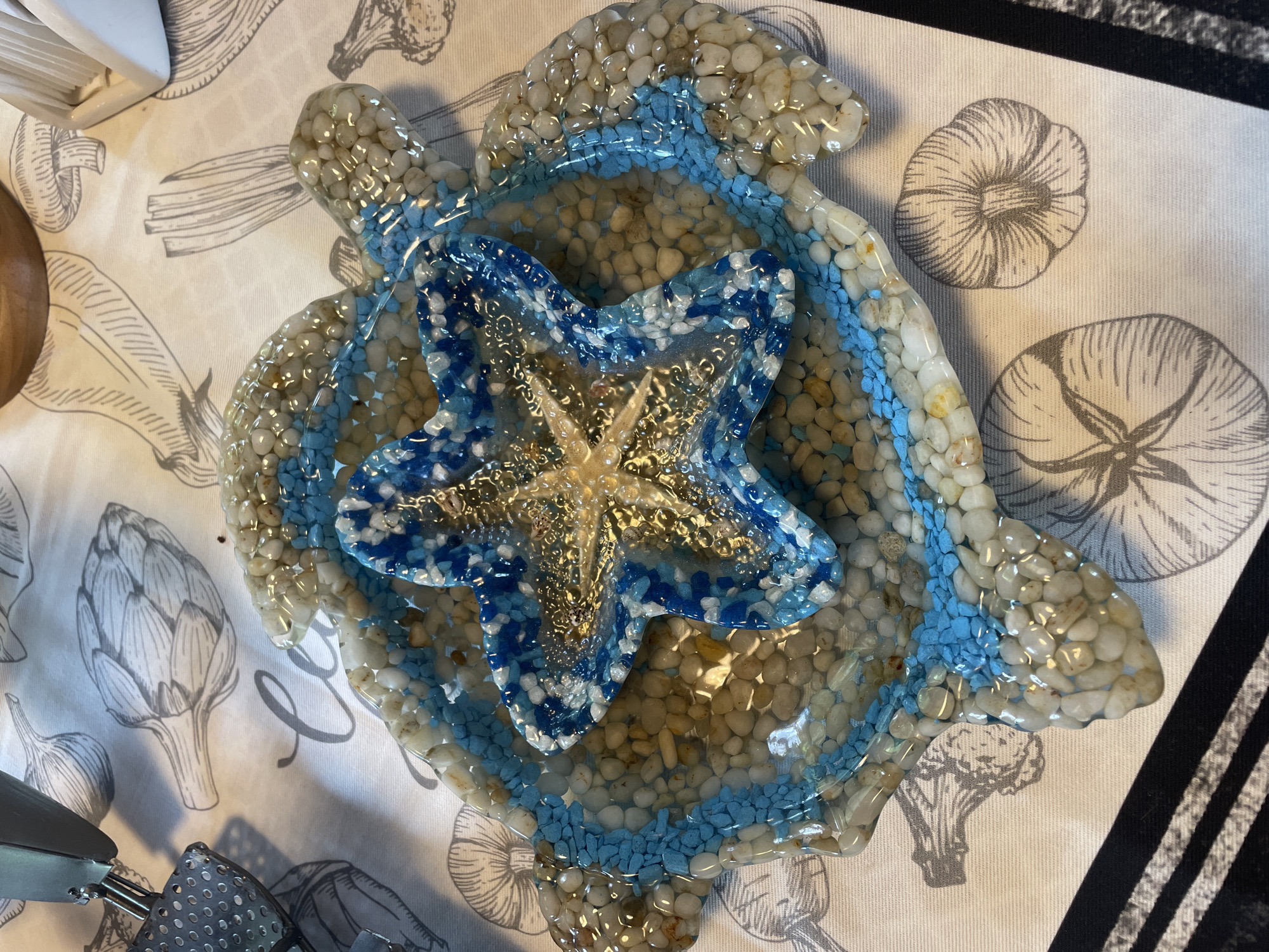 A close-up, top-down shot shows a resin art piece in the shape of a turtle, filled with small, smooth pebbles and a blue and white starfish ornament in the center. The turtle-shaped piece is on a tablecloth with a printed design of various vegetables and calligraphy. The scene is indoors, likely in a home, with no indication of the city of Турово, Russia.