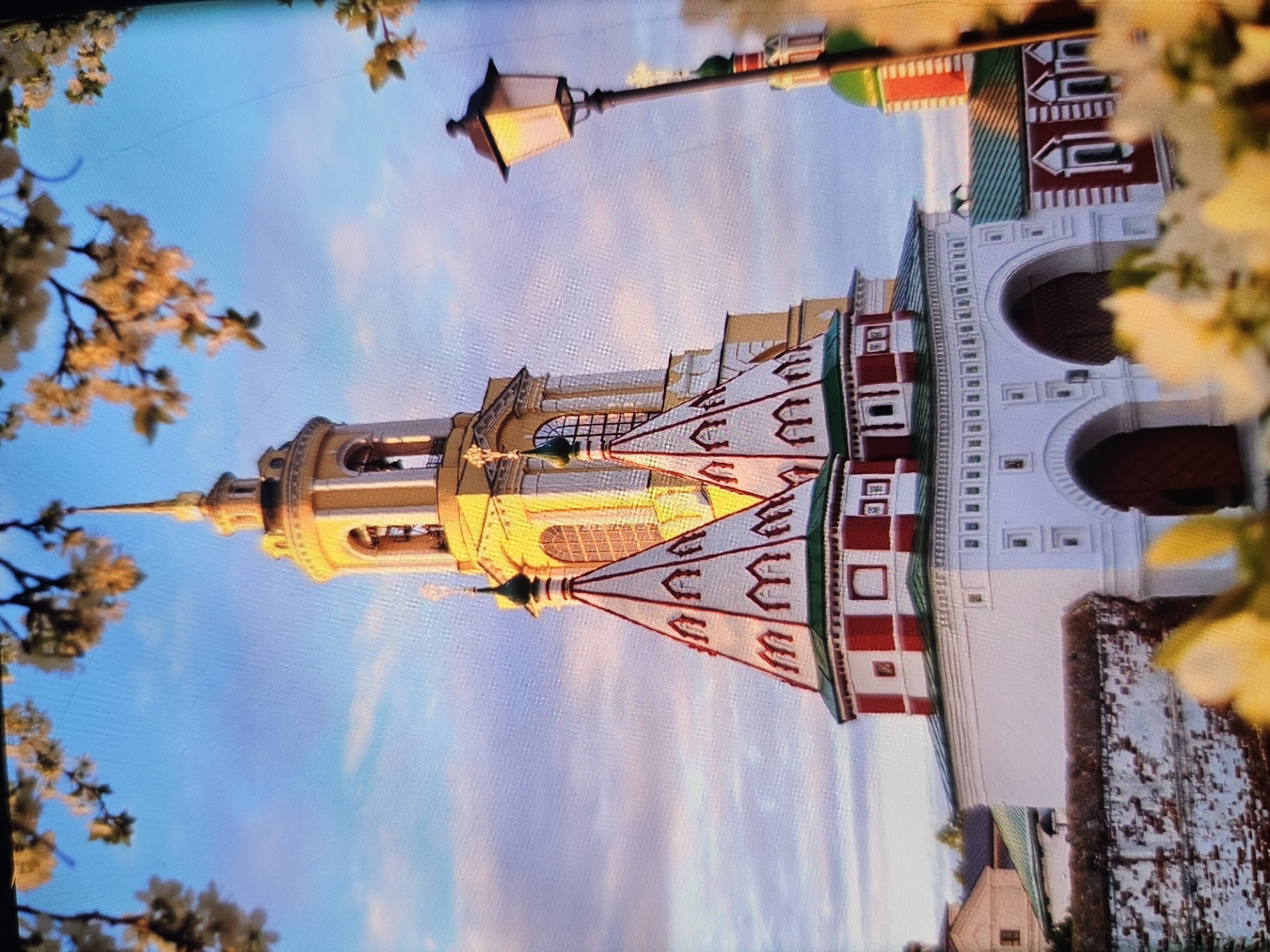 The image shows a view of the Suzdal Kremlin in Vladimir, Russia. The foreground is blurred with white and yellow flowers. Behind the flowers, a white brick wall is visible. To the right of the wall is a white arched gateway with red doors. Above the gateway, two conical red and white topped structures rise. In the background, a tall yellow bell tower with a golden spire dominates the skyline. The sky is a bright blue with wispy white clouds. A black lamppost with a glowing lantern stands to the right of the bell tower. Several other red and green topped buildings are also visible. The scene is bathed in the warm light of the setting sun.