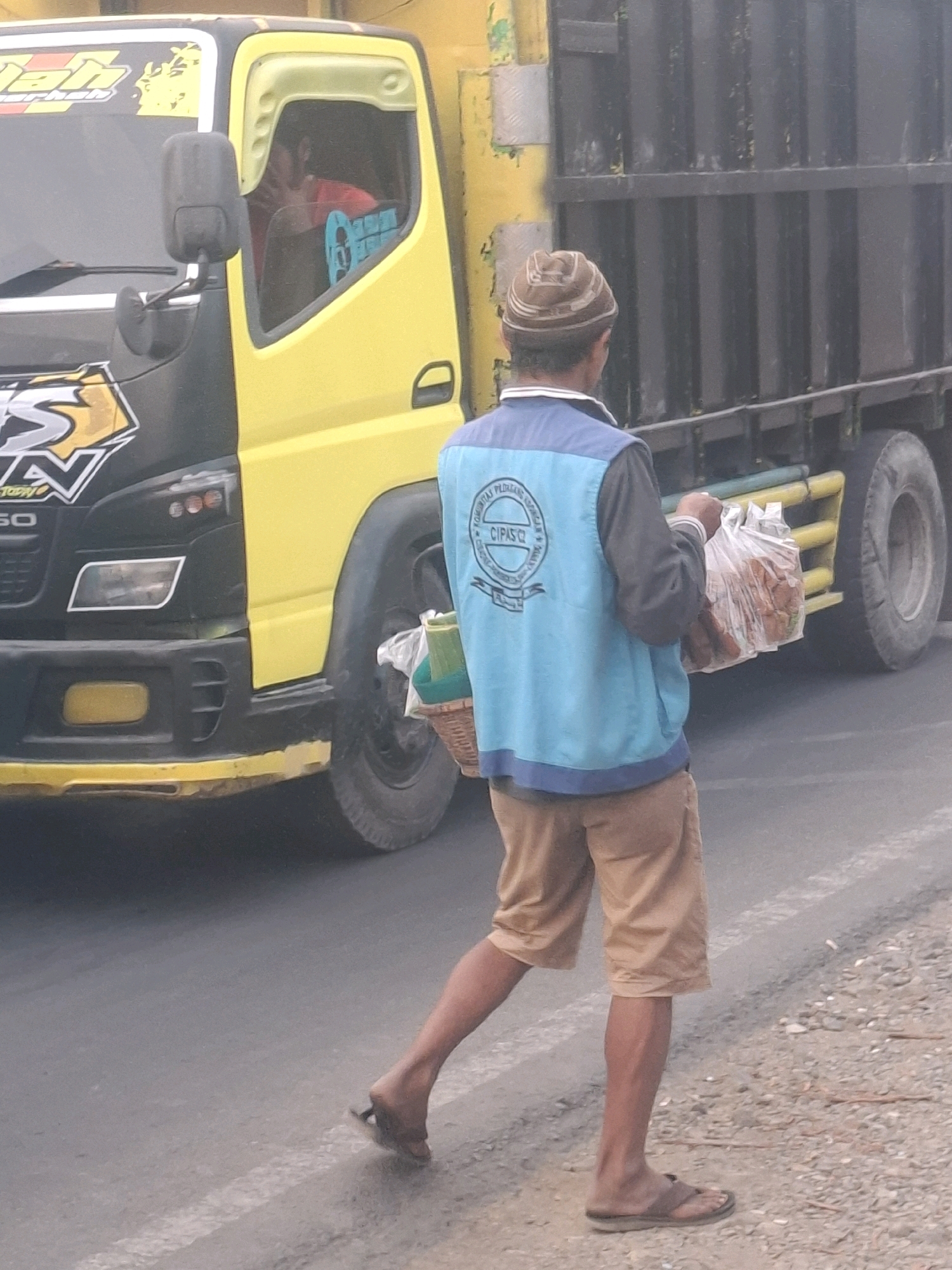 A person wearing a brown knit cap, a blue vest over a dark long-sleeved shirt, and tan shorts walks alongside a yellow and black truck on a paved road. The person carries a clear plastic bag filled with food items in their right hand and a woven basket with green items in their left hand. The truck is a commercial vehicle with a visible cargo area and the front cab appears to be yellow and black with various stickers. The environment is outdoors, suggesting a road in a potentially rural or semi-urban setting. The lighting indicates it is daytime. The image is cropped, focusing on the person and the truck, with the road surface and surrounding gravel visible.