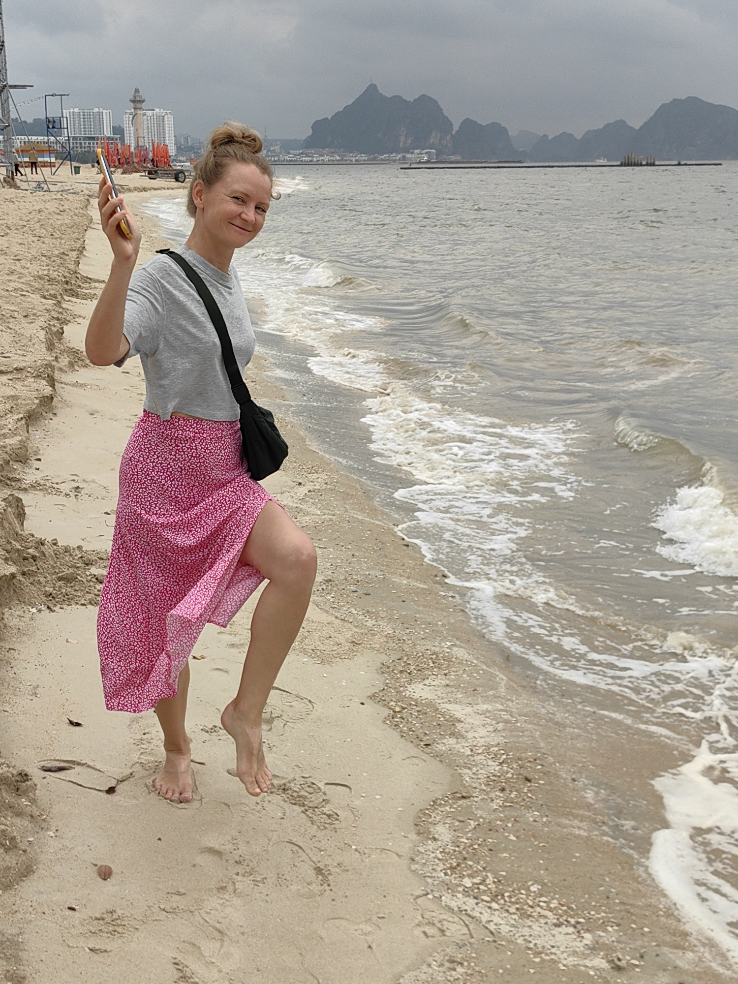 A woman with her blonde hair tied up in a bun is standing barefoot on a sandy beach in Bai Chay Ward, Vietnam. She's wearing a light grey t-shirt, a pink floral midi skirt, and a black shoulder bag. She's holding a cell phone up in her right hand. The beach is visible in the foreground with waves lapping the shore. In the background, there is a cityscape with several buildings and a coastal mountain range under a cloudy sky. A construction site can be seen to the left of the woman.