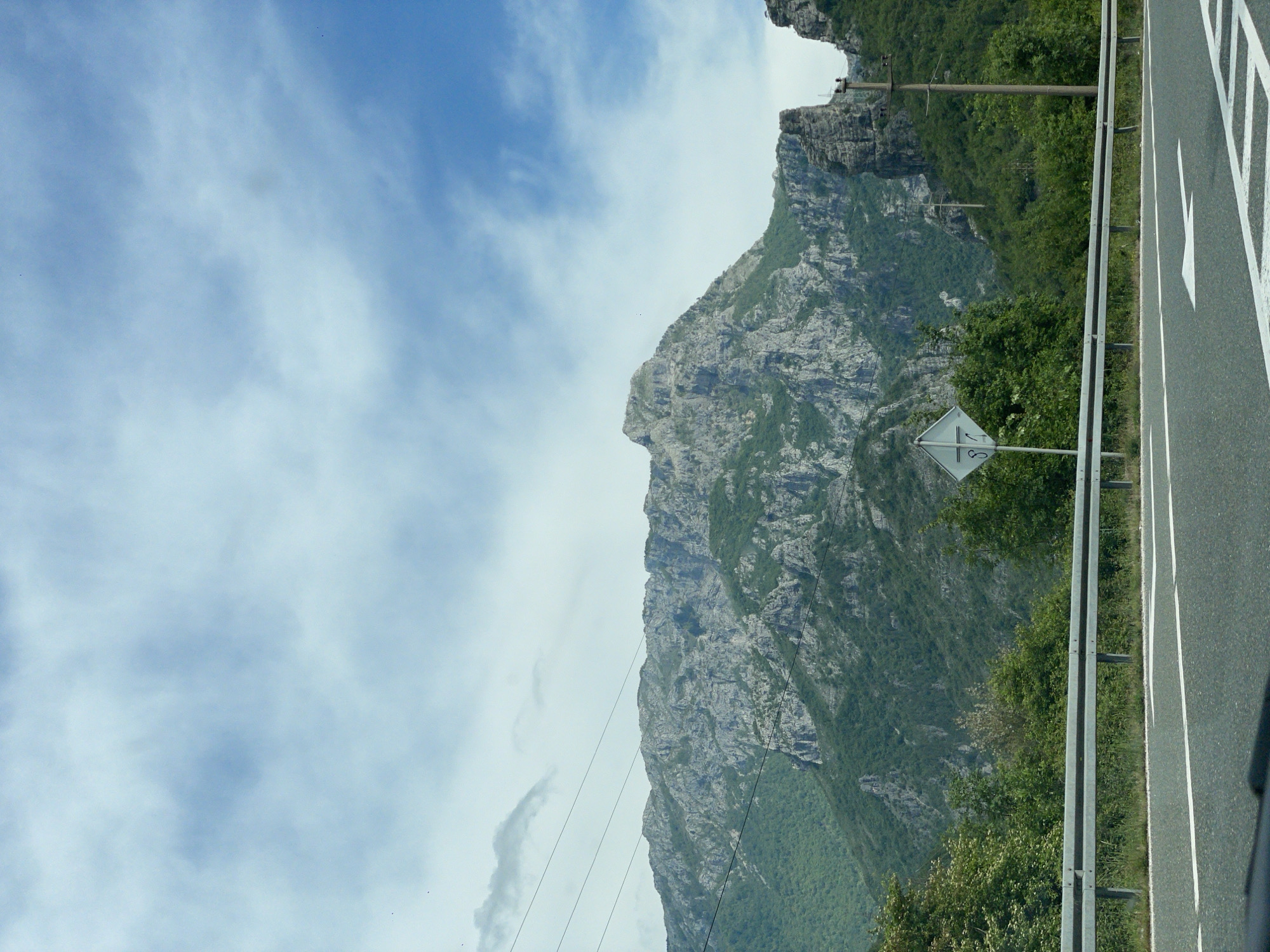 A roadside view in Donji Jasenjani, Bosnia and Herzegovina, presents a gray asphalt roadway with white directional arrows adjacent to a metal guardrail. A diamond-shaped road sign stands amidst green foliage beside the road. A towering, rugged mountain with exposed gray rock faces and patches of green vegetation dominates the scene, juxtaposed against a blue sky with scattered clouds. Overhead power lines stretch across the landscape.