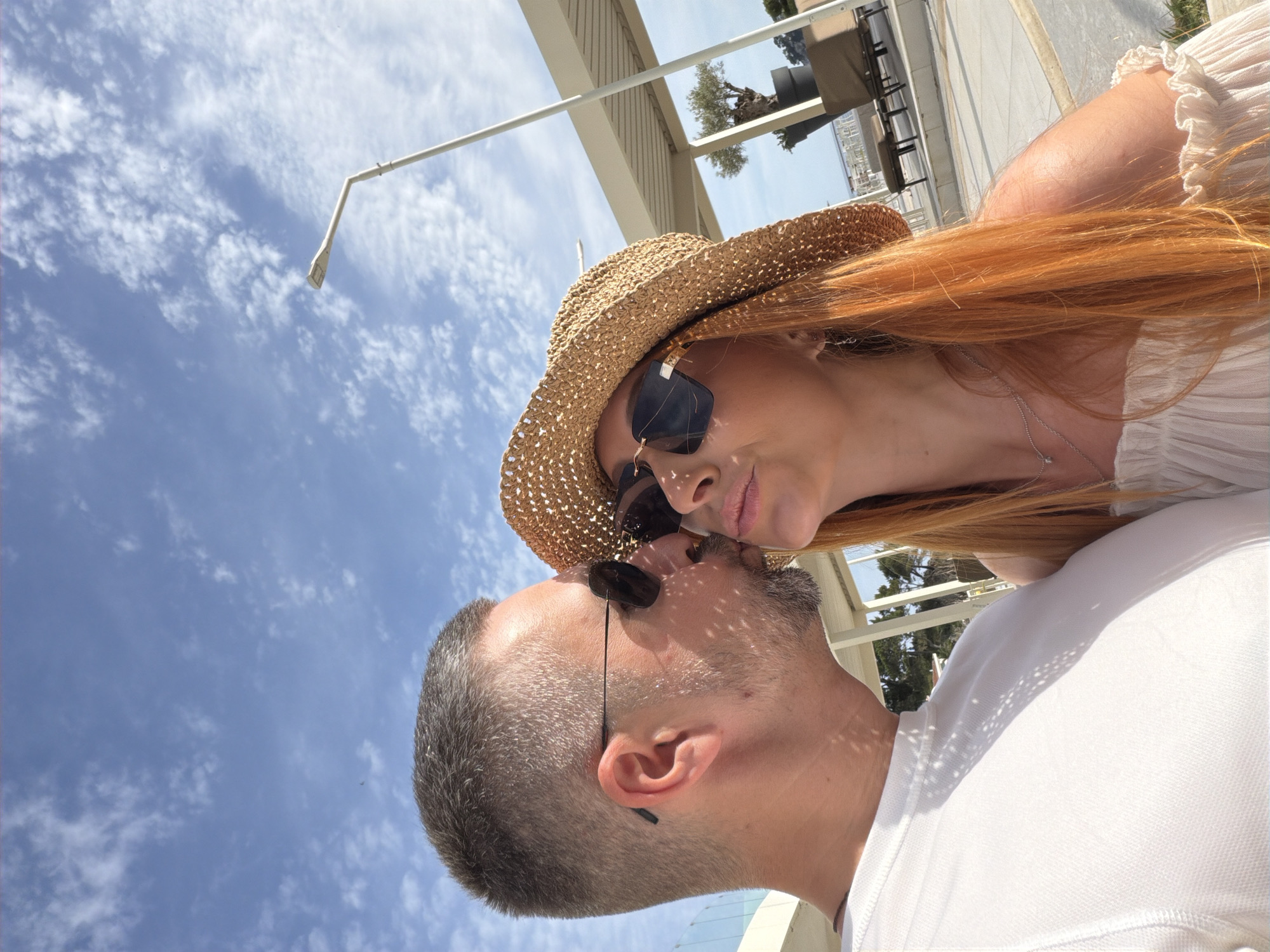 A daylight outdoor photograph captures a couple on what appears to be a resort or hotel terrace in Petrovac na Moru, Montenegro. On the left, a male with short gray hair and a gray beard, wearing dark sunglasses and a white short-sleeved shirt, is kissing the right cheek or side of the mouth of the female on the right. The female has long, reddish-auburn hair and wears a straw hat, dark sunglasses, a light-colored off-the-shoulder top, a silver necklace, and earrings. Strong sunlight casts distinct shadows, including dappled patterns from the straw hat and a structure above onto the individuals. The background features light-colored modern architectural elements, including a canopy or pergola structure and pillars. Underneath this structure, a potted olive tree and two dark lounge chairs are visible. A modern street lamp extends into the upper right portion of the frame. The sky above is clear blue with scattered white cumulus clouds. A glass railing is partially visible on the far left.