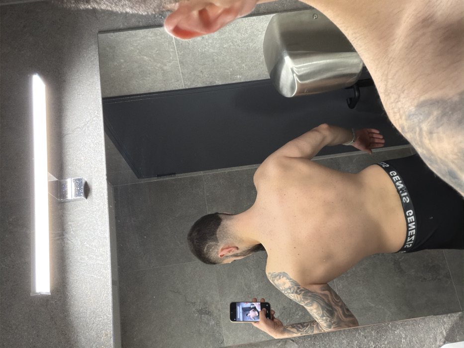 A male subject is depicted taking a mirror selfie within a modern, tiled room, identified as a facility in Plovdiv, Bulgaria. The specific location is inferred to be a Genesis Fitness & Wellness center due to the "GENESIS" branding visible on the subject's boxer briefs.
The scene captures the reflection of a male from behind, standing topless, showcasing a lean physique. He has short dark hair with a distinct shaved pattern on the nape, and a beard. His left arm is extensively tattooed, and his bare back is visible. He wears black boxer briefs with a white waistband displaying the reversed word "GENESIS". In his left hand, he holds a black smartphone, positioned to capture his reflection in the large, rectangular mirror. A smaller, vertical image of the subject's reflection is visible on the smartphone screen. The foreground shows a partial direct view of the subject's right shoulder, upper arm with tattoos, and ear, indicating he is standing very close to the mirror while photographing.
The environment features large, rectangular grey wall tiles with visible grout lines. A horizontal LED light fixture is mounted above the mirror. To the right side of the reflection, a silver, wall-mounted hand dryer is present. A dark grey door with a visible frame is also reflected in the mirror behind the subject.