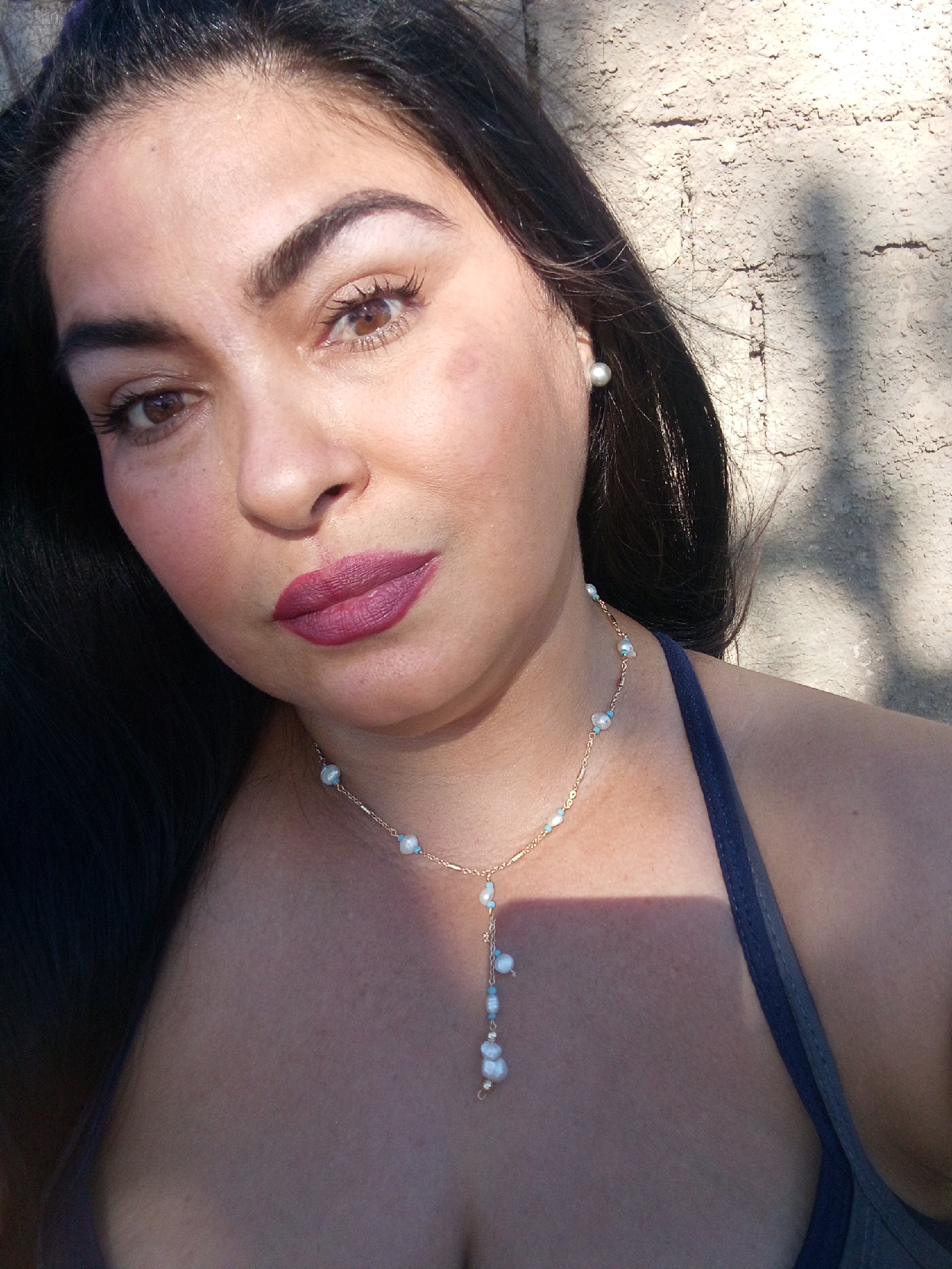 A close-up selfie captures a woman with dark hair, light skin, and brown eyes. She wears a pearl earring on her left ear and a delicate gold necklace with small white and turquoise beads. Her lips are painted a dark pink color. The background appears to be a textured wall illuminated by sunlight. The location is Juan Griego, Venezuela.