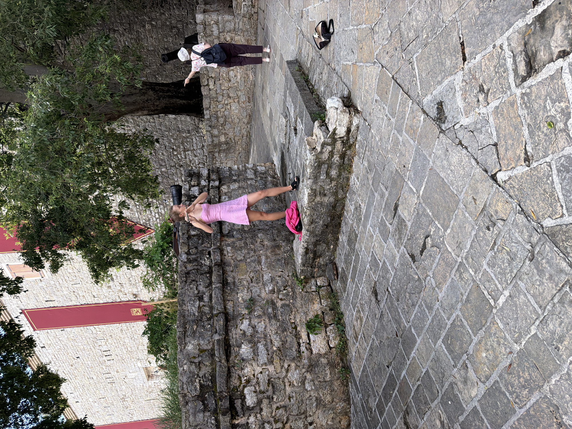 The image, when viewed upright, depicts an outdoor scene within a historic location, likely a fortified area in Budva, Montenegro, given the context. The ground is paved with irregular gray cobblestones, showing cracks and sparse vegetation.
Two women are the central subjects. In the mid-ground, a young woman with light brown hair, wearing a light pink dress and black shoes, stands beside a stone wall. She holds a device, possibly a phone or small camera, near her face, suggesting she is taking a photo or selfie. A bright pink bag rests on a stone ledge next to her. Further back and to the right, an older woman, viewed from behind, wears dark pants, a patterned shirt, a white cap, and a dark backpack. She is pointing with her right hand towards the left, possibly indicating a point of interest. A black bag and what appear to be sandals are on the cobblestones near her.
The setting is dominated by ancient stone architecture. A prominent, rugged stone wall runs along the left side of the scene, behind the younger woman. Lush green trees with dense foliage are visible behind and above this wall. In the background, a large, light-colored stone building or wall is decorated with several tall, vertical red banners. These banners have gold borders and feature a crest or symbol near the top, although no specific text is clearly legible. The scene is brightly lit, indicating a sunny daytime. The overall impression is that of tourists exploring a historical site.