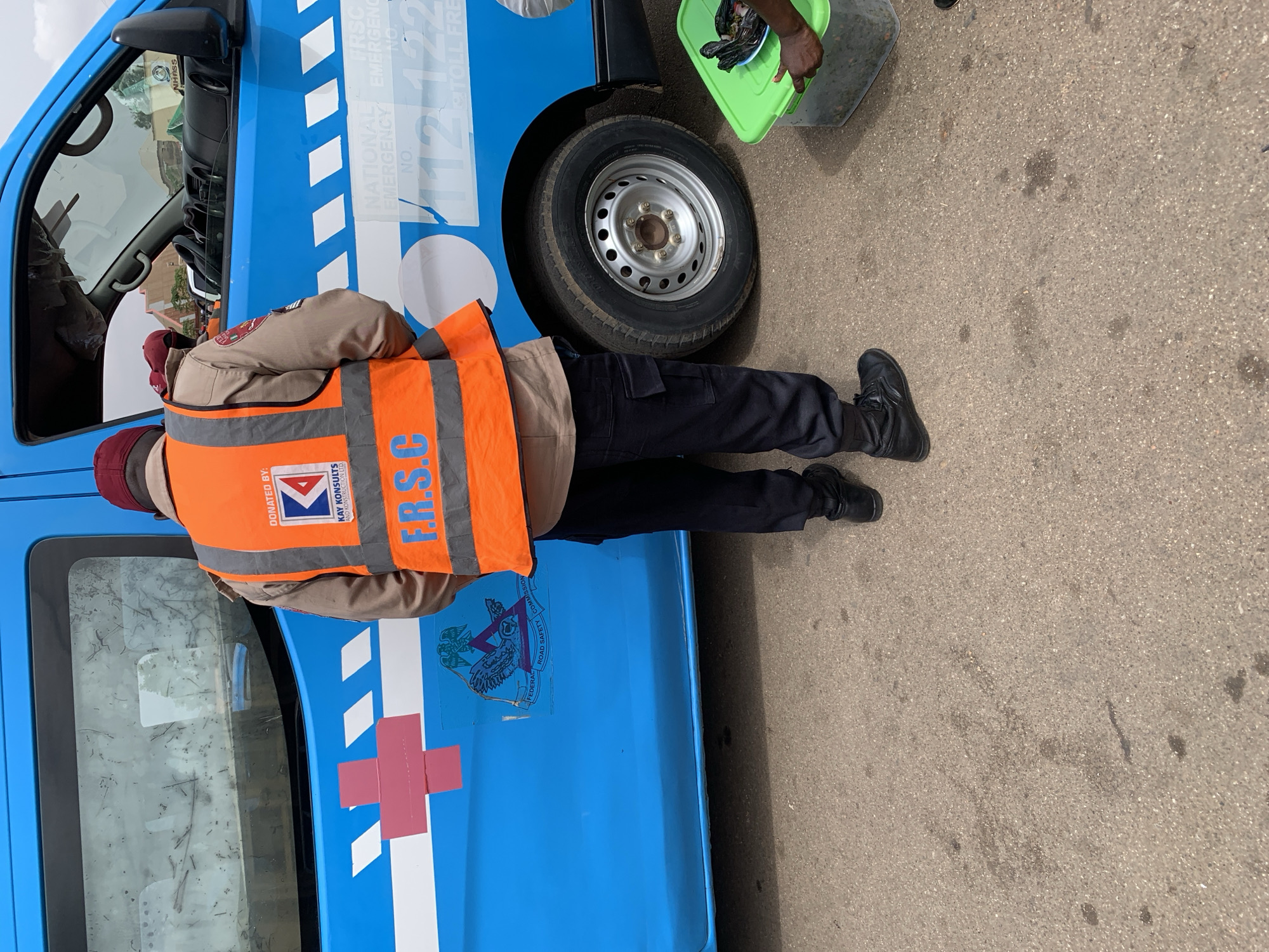 The image shows a person in a Federal Road Safety Commission (FRSC) uniform, consisting of black trousers, black shoes, a khaki shirt, and an orange reflective vest with "F.R.S.C" printed on it. They are standing next to a blue FRSC emergency vehicle. The vehicle has a white cross on the side and FRSC emergency contact information printed on it. Another person's hand is visible, holding a green container. The scene appears to be on a paved road or street. According to the FRSC's official website, they have offices and patrol operations throughout Nigeria. Without more information, it is difficult to confirm this image was taken in Bukuru.