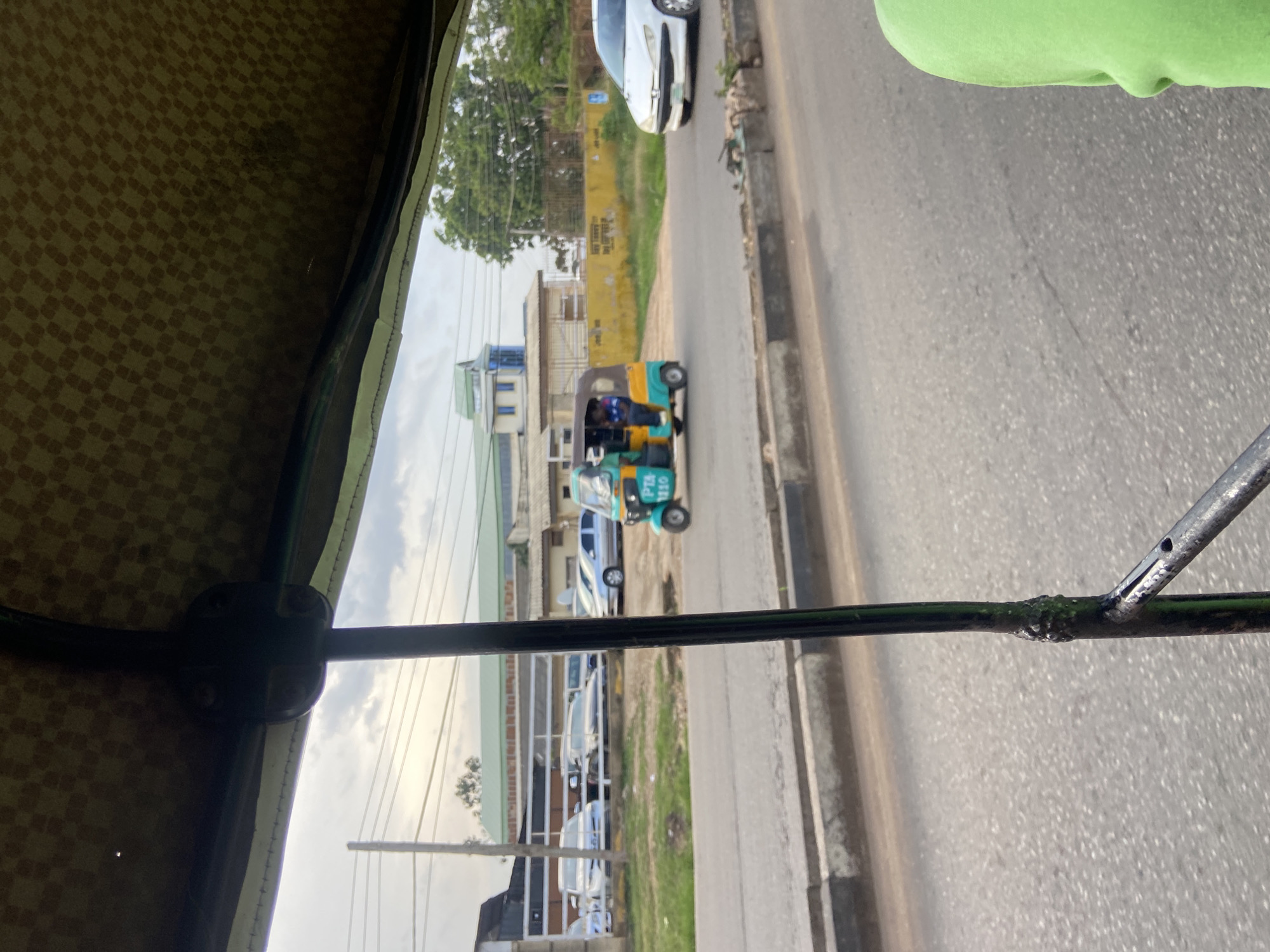The image, taken from inside a vehicle, shows a street scene in Jos, Nigeria. A yellow and blue auto-rickshaw, a common mode of transport in the city, is visible on the road. The auto-rickshaw has license plates indicating "PTA 1110". Several cars are parked along the side of the road, and a white car is driving in the opposite direction. In the background, there is a building with a green roof and blue accents, and some trees. The sky is cloudy. The foreground shows part of the interior of the vehicle from which the photo is taken, including a black metal bar and a patterned canopy overhead. The road appears to be asphalt, with a concrete curb.