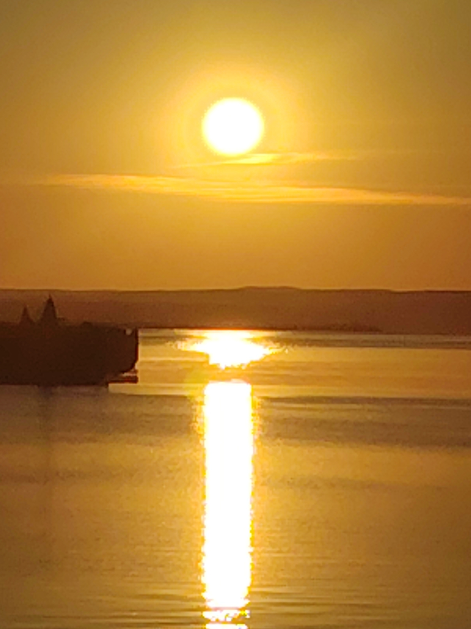 The image depicts a serene sunset over a body of water, likely the Kama River, in Chaikovsky, Russia. The sun, a bright orb of light, is positioned high in the sky, casting a warm, golden glow over the scene. A thin band of clouds stretches horizontally across the horizon. In the distance, silhouetted against the sunset, is the faint outline of buildings, possibly part of the Chaikovsky landscape. The water reflects the sun's brilliance, creating a shimmering path of light that extends towards the foreground. The overall impression is one of tranquility and natural beauty.