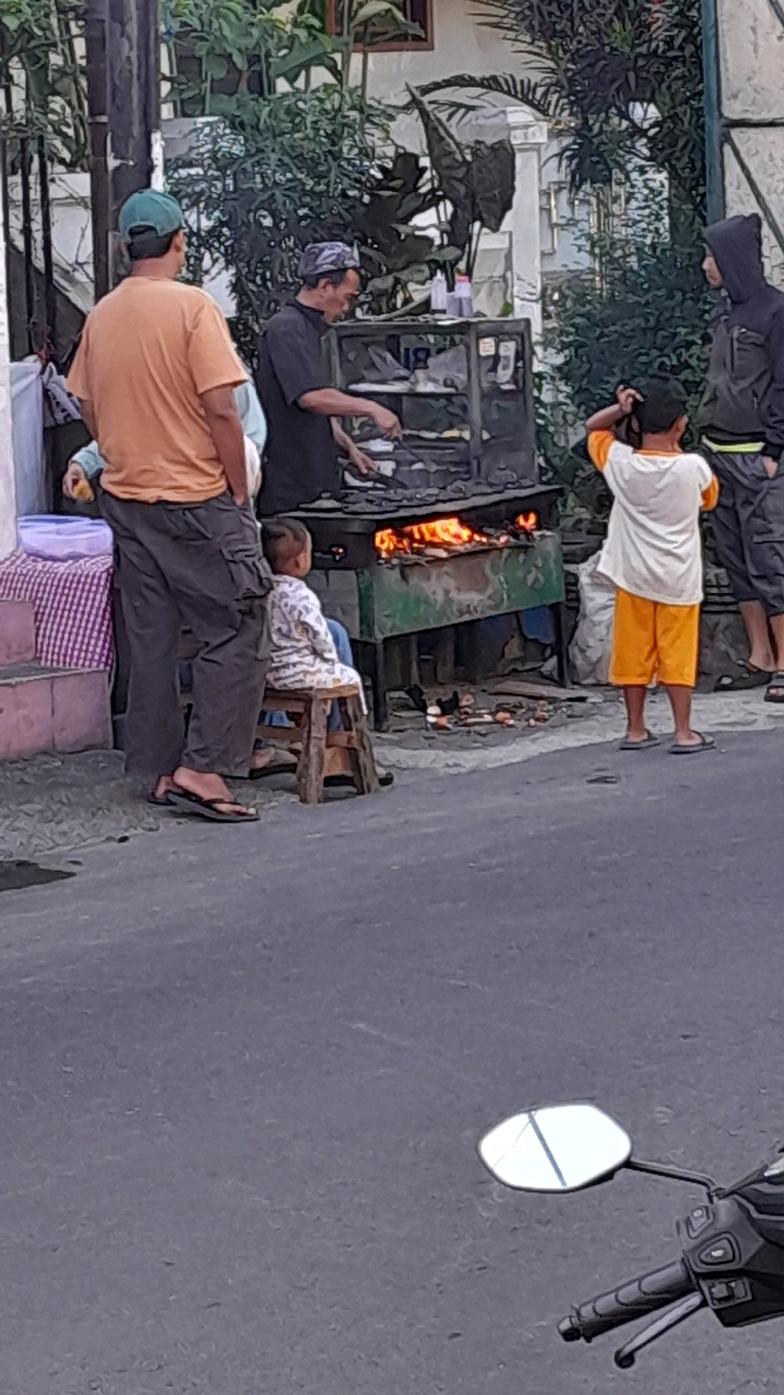 The image depicts a street scene in Bandung City, Indonesia, where a man is cooking food over a grill. He is wearing a black shirt and a headscarf. In front of him is a food cart with a glass display and a burning grill. A young child, wearing a white and patterned shirt and blue shorts, is sitting on a wooden stool to the left of the grill, facing it. To the right of the grill, a boy in an orange shirt and shorts stands looking at the scene. Behind the boy, another person in a black hooded jacket is standing. A man in an orange t-shirt and dark pants is standing to the left of the food cart, facing the grill. The background shows greenery and buildings. A motorcycle mirror is visible in the foreground.
