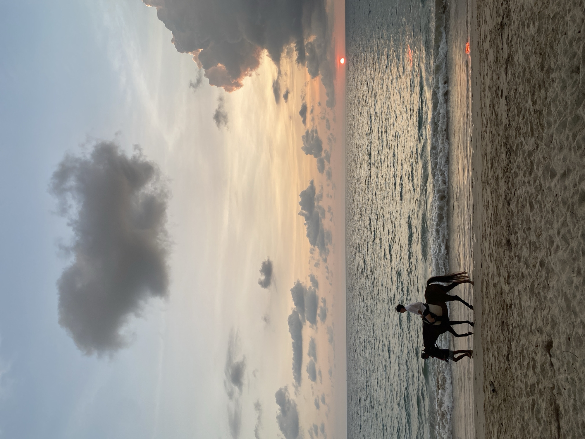 This image captures a serene sunset scene on a beach in Choeng Thale, Thailand. The sun, a vivid orange sphere, is visible low on the horizon, casting warm light across the sky and water. The sky transitions from a soft blue at the top left to brilliant oranges and yellows closer to the horizon, dotted with scattered clouds. Some clouds appear dark and grey, while others are illuminated with golden edges by the setting sun.
The right side of the image is dominated by the ocean, with gentle waves lapping at the shore. The water's surface shimmers, reflecting the intense colors of the sunset. The foreground and bottom left show the sandy beach, textured and damp where the waves recede.
In the shallow water near the shoreline, a dark-colored horse stands, carrying a rider. The rider, wearing a light-colored shirt, is silhouetted against the bright reflection on the water. Another individual, also mostly in silhouette, stands beside the horse in the water, appearing to guide or attend to it. The scene depicts a tranquil moment of horseback riding on the beach, likely a recreational activity enjoying the beautiful end-of-day light. There is no visible text in the image.