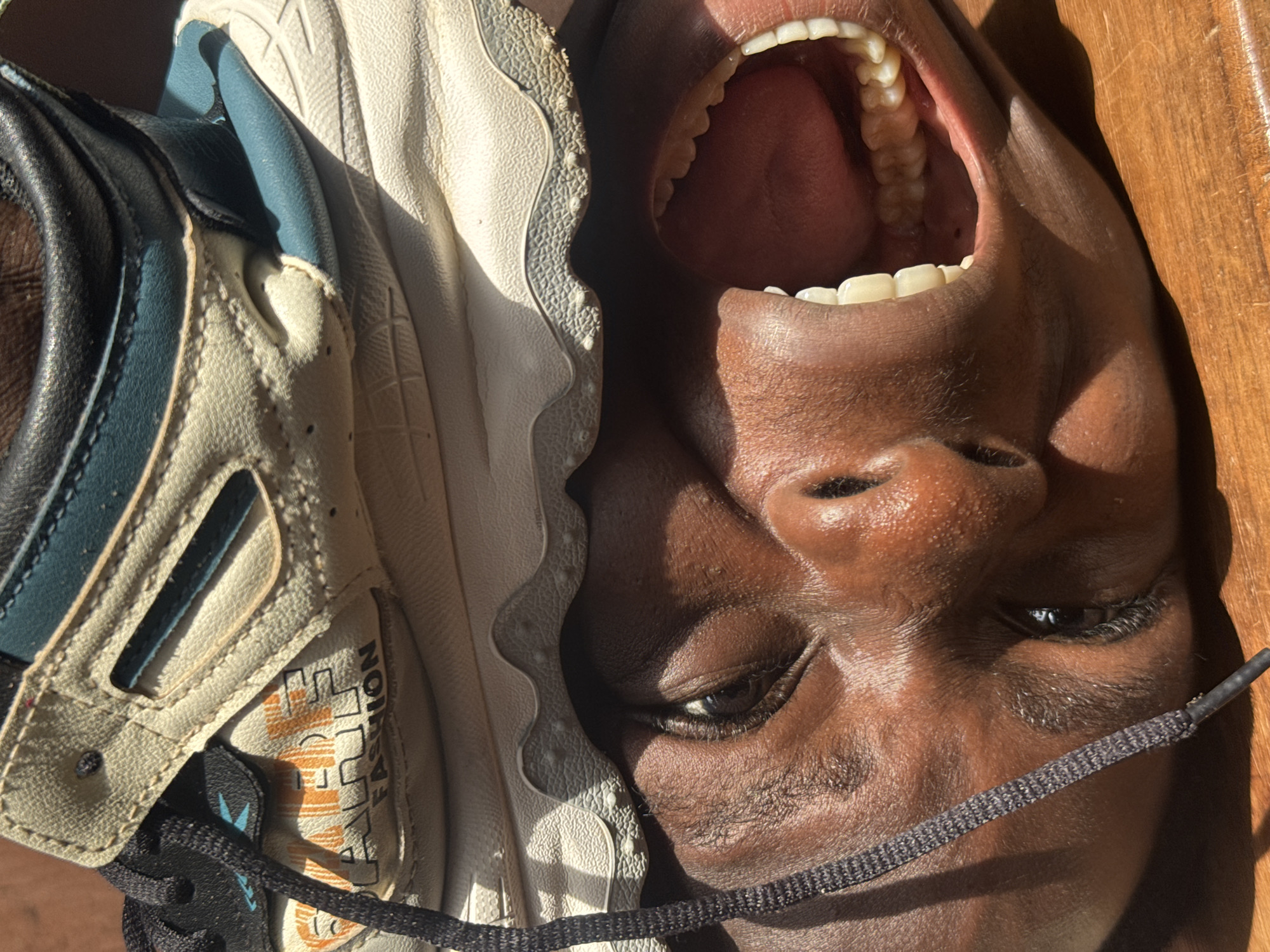 The image shows a close-up of a person's face juxtaposed with a sneaker. The person's face is tilted, with their mouth open showing teeth and tongue. The face is dark-skinned, with visible details like eyelashes and pores. The sneaker, partially obscuring the face, is a light color with dark accents and the word "FASHION" printed on it. A shoelace lies across the face. The background is a mix of shadows and light, including the texture of a wooden surface. There is no location information related to Onitsha, Nigeria.