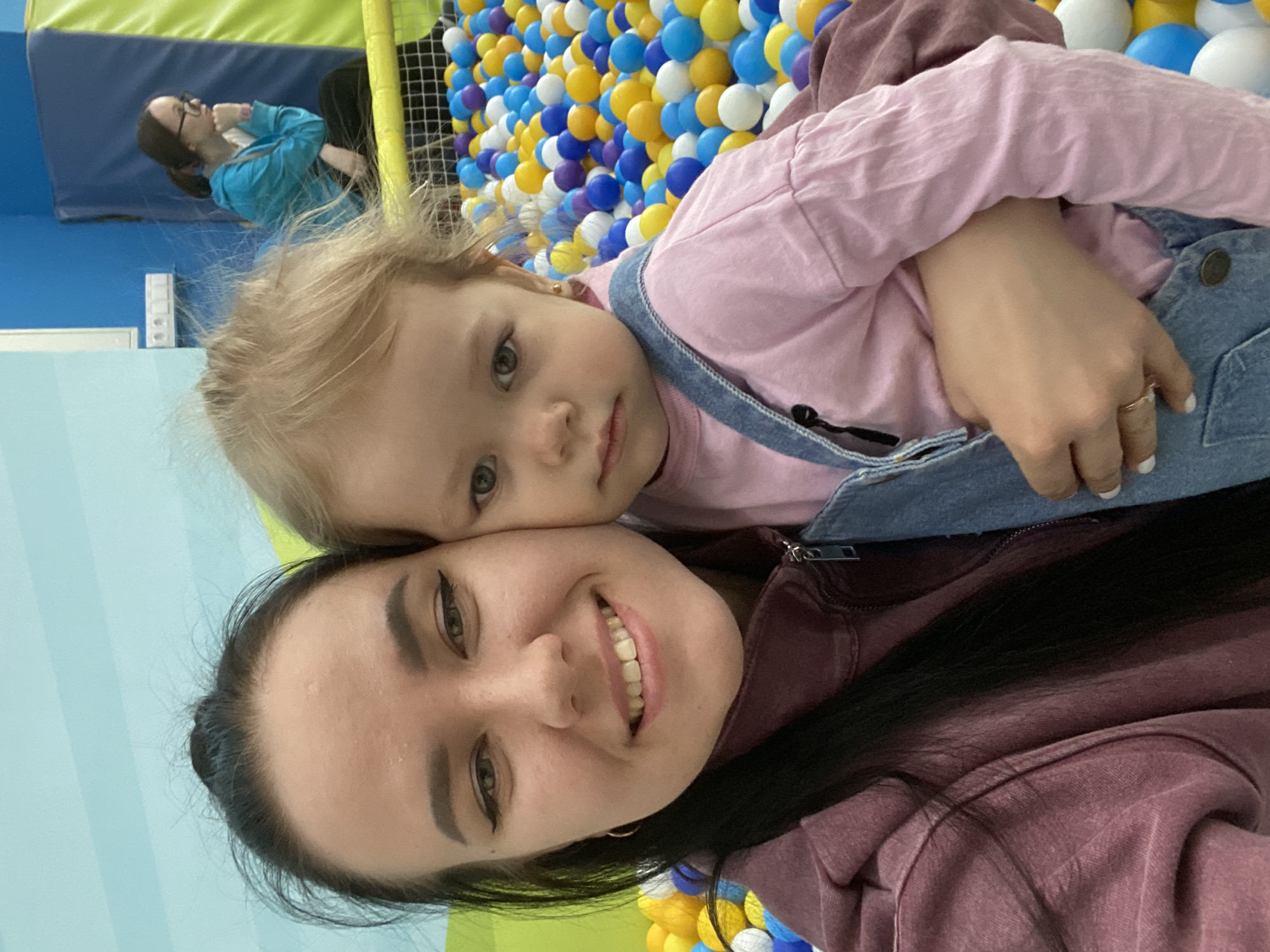 A selfie photograph shows a woman holding a toddler against a backdrop of a ball pit in an indoor play area. The woman has fair skin, dark eyebrows, and dark hair pulled back. She is wearing a maroon-colored hooded sweatshirt. The toddler is fair-skinned with blonde hair and is wearing a pink long-sleeved shirt and denim overalls. The ball pit is filled with yellow, blue, purple, and white plastic balls. In the background, partially obscured, a woman with glasses can be seen in the play area. The location is likely in Perm, Russia.
