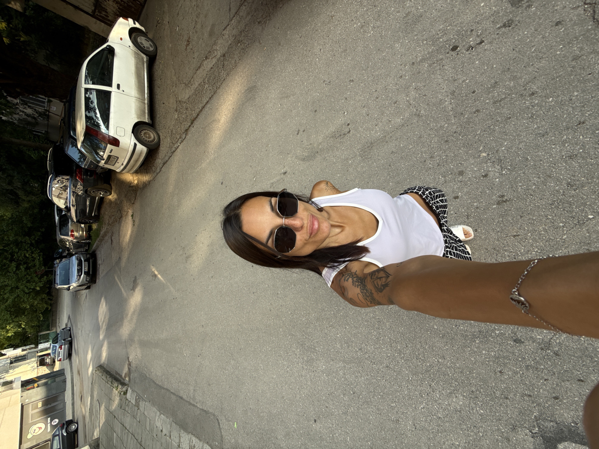 In the city of Pleven, Bulgaria, a woman is captured in a high-angle selfie on a paved street. She is wearing a white tank top, black and white patterned pants, white sandals, and large sunglasses. Her left arm is adorned with tattoos, and a silver bracelet is visible on her right wrist. The background shows parked cars, including a white Fiat Punto, and buildings, some with trees in front of them. The lighting suggests it is late afternoon or early evening.