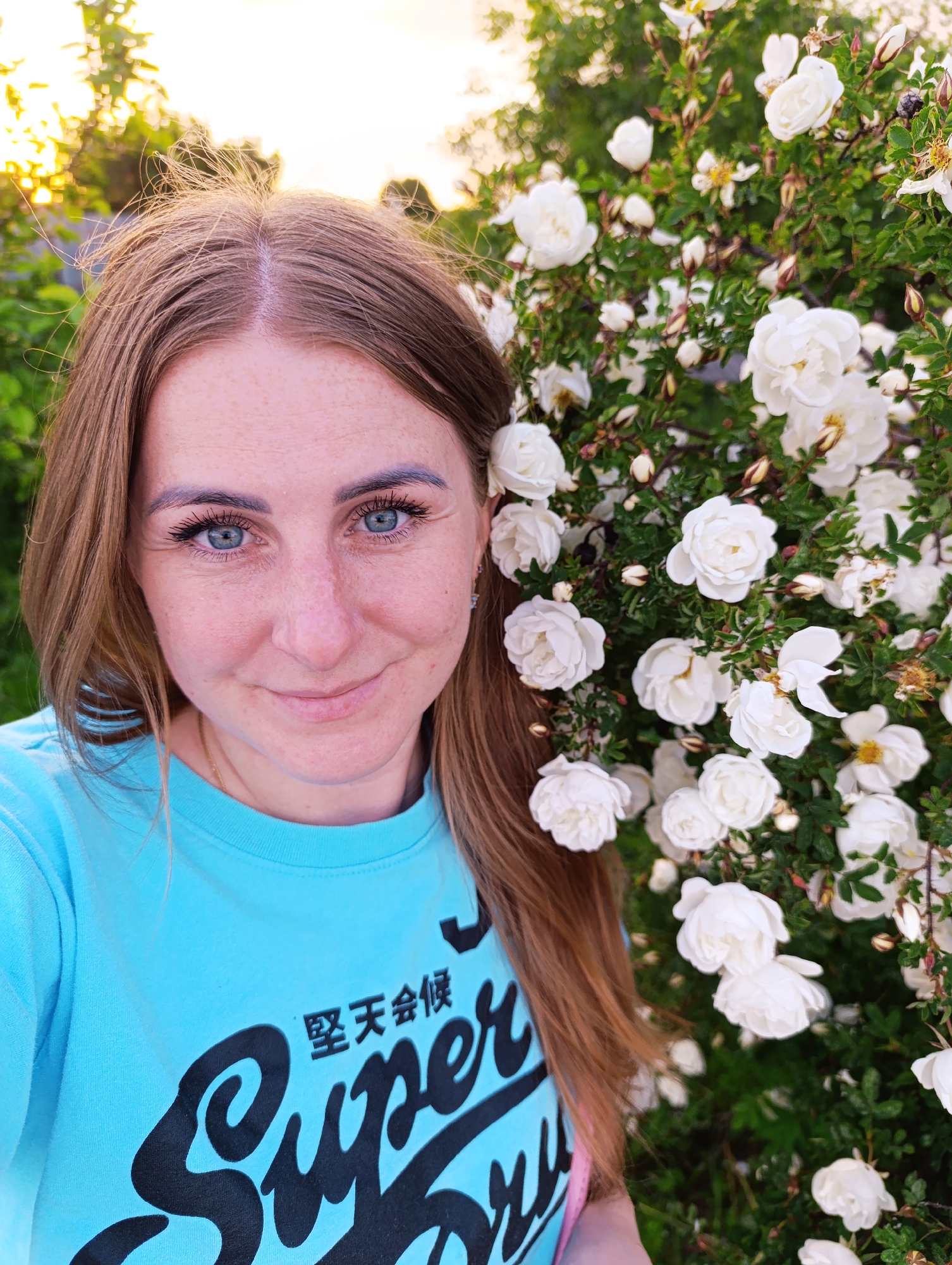 A close-up outdoor portrait captures a light-skinned woman with long, reddish-brown hair and blue eyes, looking directly at the camera with a soft, friendly smile. Freckles are visible across her nose and cheeks. She wears a bright turquoise or aqua long-sleeved t-shirt, on which black, stylized text is visible. The text includes Japanese characters "堅天会候" above the partially visible English word "Super." A subtle gold chain necklace can be seen around her neck.
She is positioned to the left of the frame, with her head and upper torso filling much of the left side. To her right, a dense rose bush in full bloom occupies the background. The bush is laden with numerous small, white roses, some fully open, others as buds, and a few with spent petals. The flowers and green foliage create a vibrant backdrop.
The lighting suggests late afternoon or early evening, with a warm, golden glow emanating from the upper left background, indicating a setting sun or golden hour. This warm light illuminates her hair, particularly around the crown and part, highlighting individual strands. The overall mood of the image is serene and pleasant, with the woman appearing calm and approachable. The setting is likely a garden or outdoor space.