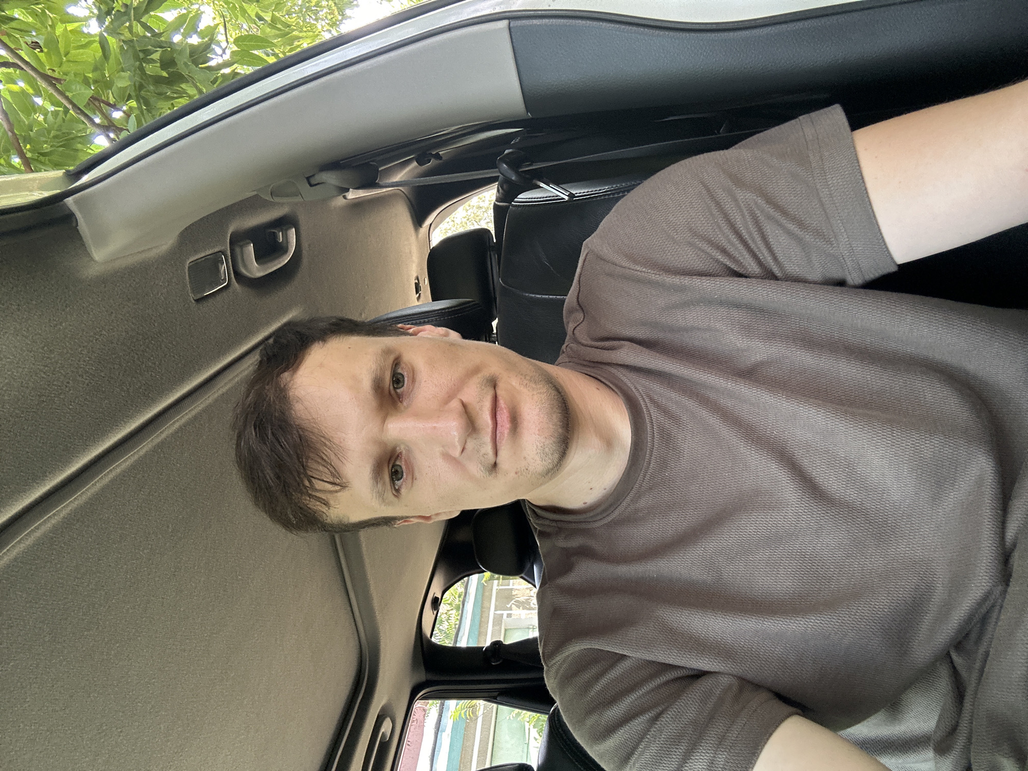 A man with light brown hair and green eyes is sitting in the driver's seat of a car. He is wearing a brown t-shirt with a textured pattern. The car's interior is visible, including black leather seats and the sunroof. Outside the car, green leaves and branches of a tree are seen. The man is looking at the camera, and his expression is neutral. The scene appears to be outdoors, possibly in a residential area, given the glimpses of buildings in the background. The image was taken in Makiv, Ukraine.