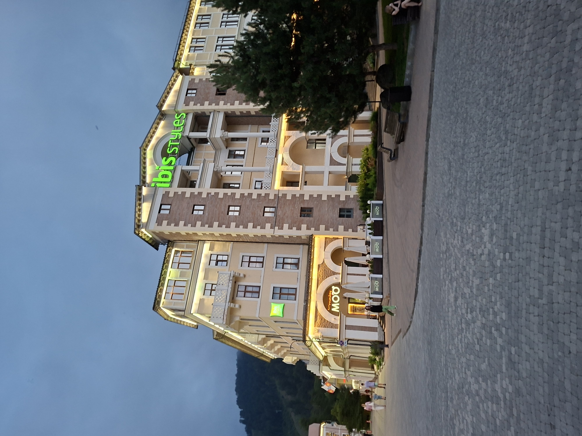 The image depicts the Ibis Styles hotel in Estosadok, Russia, at dusk. The hotel is a multi-story building with light-colored facade and brick accents. The "ibis Styles" logo is prominently displayed in green neon letters on the top of the building. Several windows are visible, some with balconies. The ground level features an arched entrance with the "monkey MOO" restaurant sign. White umbrellas suggest outdoor seating for the restaurant. In front of the hotel, a paved walkway is visible, with people strolling and sitting on benches. Trees are present on the right side of the image. The background shows a forested mountain slope, indicating a mountainous or resort area. The sky is a dark blue, characteristic of twilight.